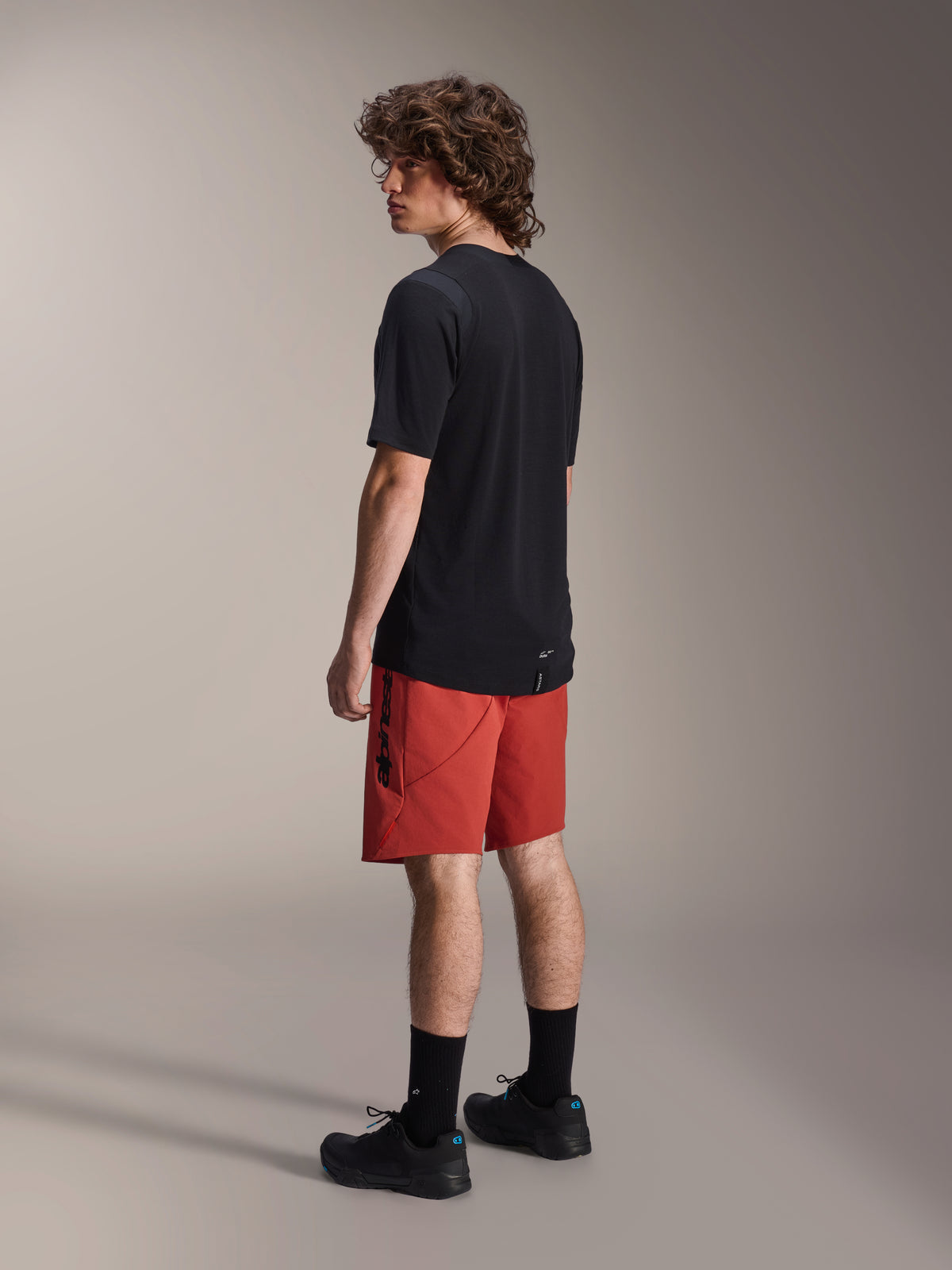 Alpinestars A-Dura Elite Shorts, spodenki rowerowe MTB, ciemnopomarańczowe, noszone z czarną koszulką z krótkim rękawem i czarnymi butami MTB, widok modelu z tyłu i boku podkreślający sportowy krój oraz branding