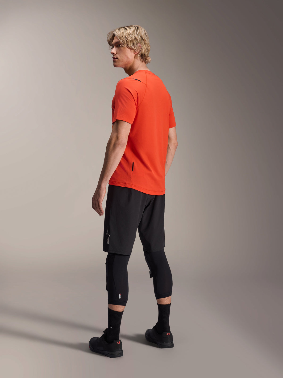 Alpinestars A-Aria Astar Short Sleeve Jersey, Spicy Orange, back three-quarter view on model looking over shoulder, paired with black MTB shorts and knee protectors, performance mountain bike apparel