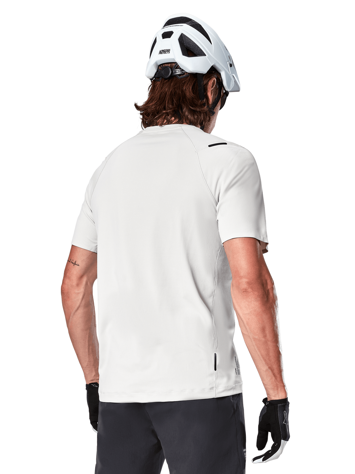 Alpinestars A-Aria Astar Jersey, krótki rękaw, kolor White Dove, widok z tyłu lekkiej i oddychającej koszulki rowerowej MTB, noszonej z białym kaskiem i czarnymi rękawiczkami rowerowymi, z wydłużonym tyłem dla lepszej osłony