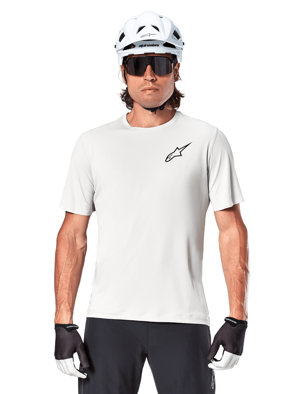 Rowerzysta w koszulce Alpinestars A-Aria Astar z krótkim rękawem, kolor White Dove z małym czarnym logo Astar na piersi, biały kask rowerowy, czarne okulary przeciwsłoneczne i czarno-białe rękawiczki, profesjonalna odzież MTB do jazdy w terenie