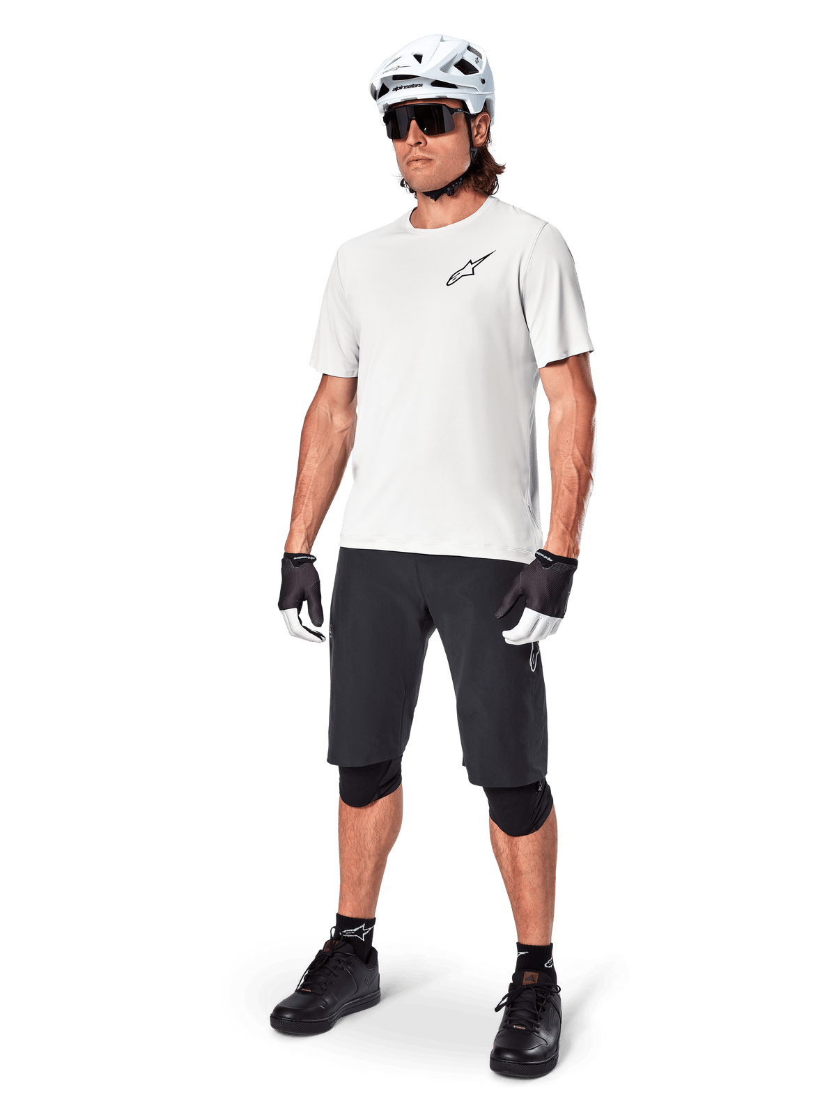 Rowerzysta MTB w koszulce Alpinestars A-Aria Astar z krótkim rękawem w kolorze White Dove, czarne szorty rowerowe, biały kask i czarne ochraniacze na kolana, małe czarne logo na piersi, dopasowany zestaw odzieży rowerowej do jazdy po szlakach