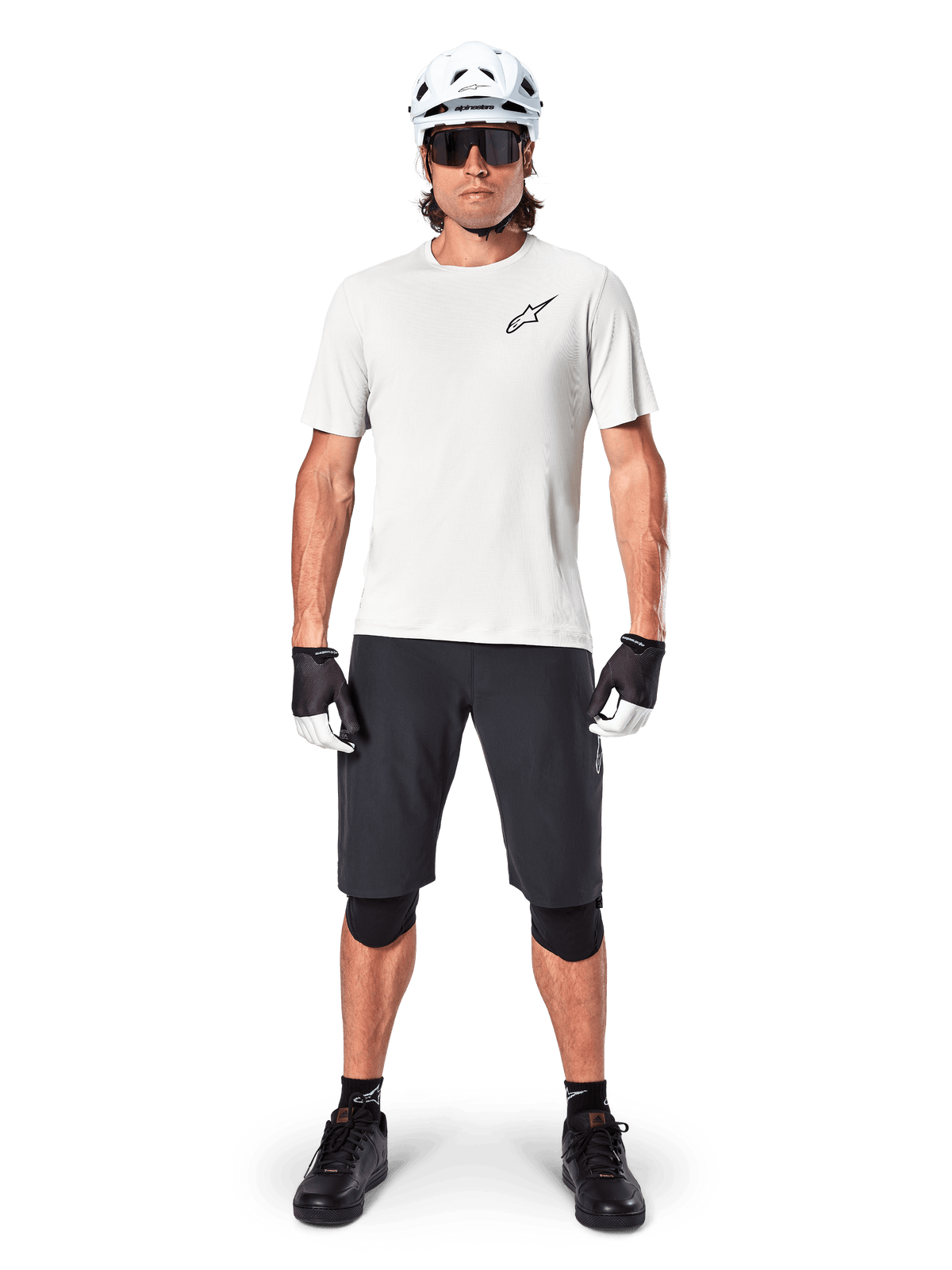 Rowerzysta ubrany w koszulkę Alpinestars A-Aria Astar z krótkim rękawem w kolorze White Dove, czarne logo Astars na piersi, czarne szorty MTB, biały kask, okulary przeciwsłoneczne, czarne ochraniacze kolan i rękawiczki, kompletny zestaw do jazdy w terenie.