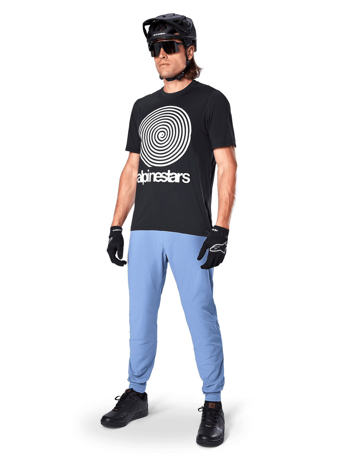 Alpinestars A-Dura Oscar Short Sleeve Jersey, Black MTB jersey featuring a large white heritage spiral graphic, worn by a rider with a black helmet, sunglasses, black gloves, and light blue trousers, performance mountain bike gear