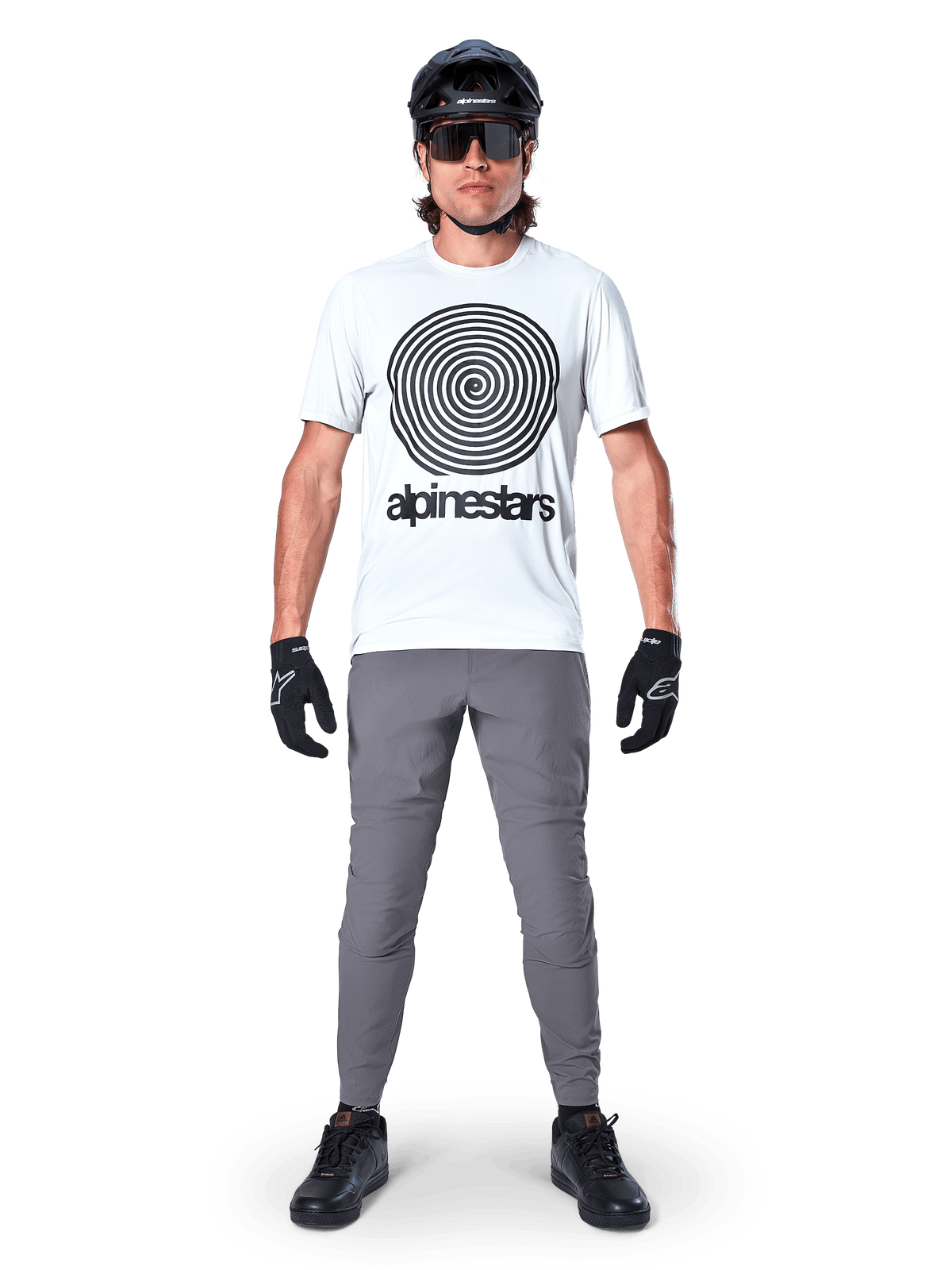 Rowerzysta górski w koszulce Alpinestars A-Dura Oscar z krótkim rękawem, kolor White Dove z dużym czarnym spiralnym logo, szare spodnie, czarny kask, okulary przeciwsłoneczne i czarne rękawiczki, wyczynowy strój MTB do jazdy trailowej