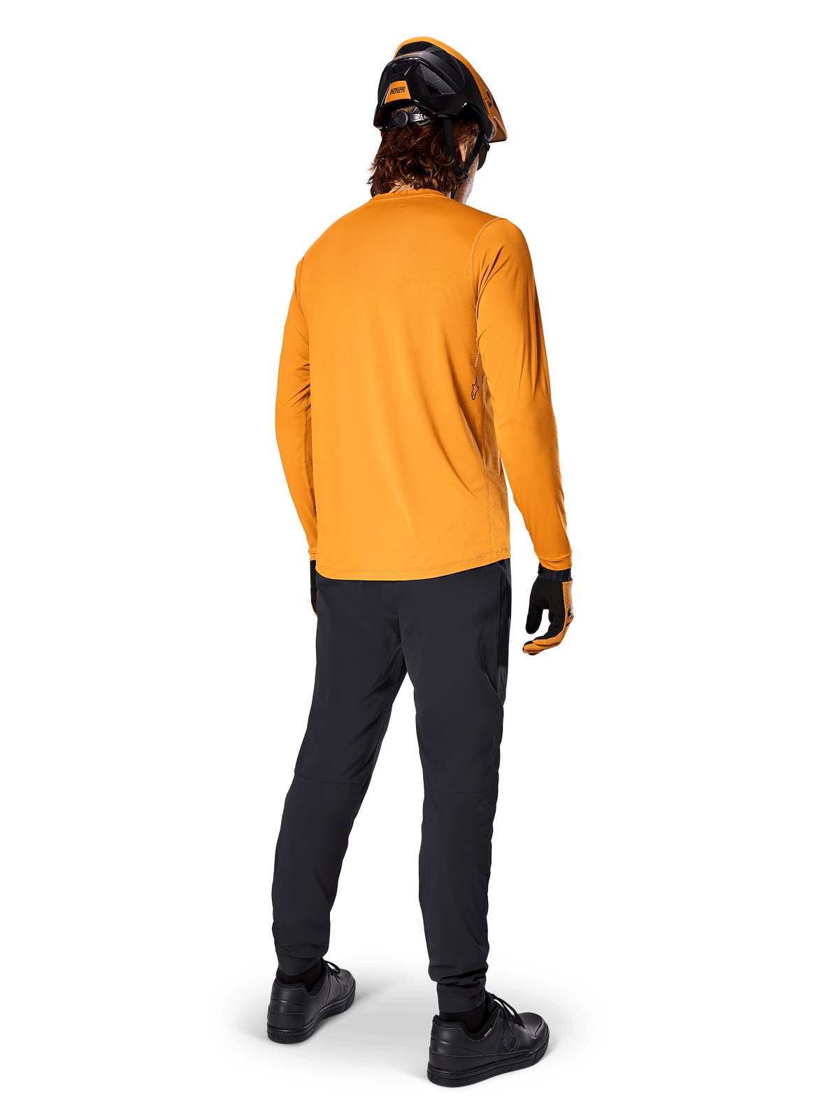 Rowerzysta górski widziany od tyłu, ubrany w koszulkę Alpinestars A-Dura Rocker Long Sleeve w kolorze Dark Gold, czarne spodnie, pomarańczowo-czarny kask i czarne buty, profesjonalny zestaw odzieży MTB do jazdy po szlakach
