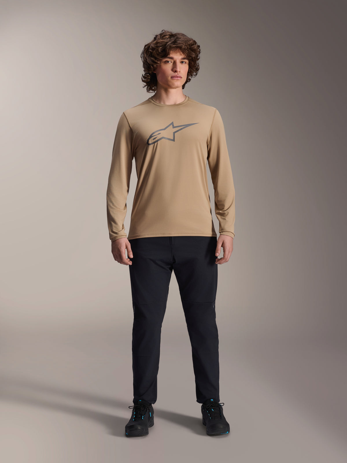 Alpinestars A-Dura Astar Long Sleeve Jersey, jasnobrązowa koszulka z długim rękawem z szarym logo Astars na piersi, noszona przez modela w czarnych spodniach technicznych i butach, casualowa odzież wyczynowa MTB