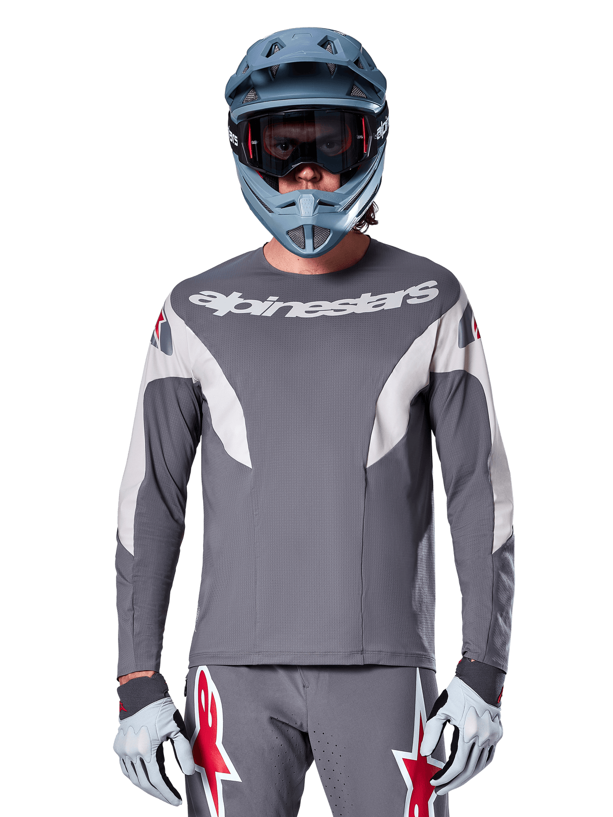 Rowerzysta MTB w koszulce Alpinestars A-Supra Race Jersey - Long Sleeve w kolorze ciemnoszarym, koszulka rowerowa z długim rękawem i białym logo Alpinestars, niebiesko-szary kask full-face, gogle i szare rękawice wyścigowe, kompletny strój wyścigowy do jazdy w terenie