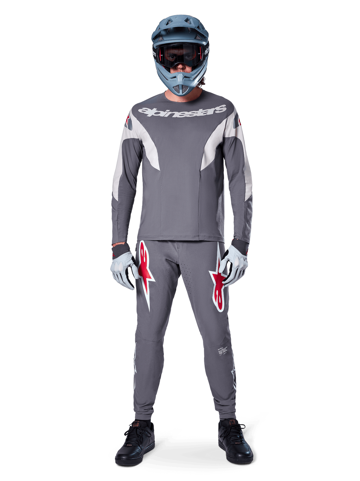 Rowerzysta w koszulce Alpinestars A-Supra Race Jersey z długim rękawem w kolorze ciemnoszarym, zestawionej z dopasowanymi szarymi spodniami z wyrazistymi czerwonymi logotypami gwiazdy, szarym kaskiem z goglami i jasnoszarymi rękawicami; kompletny zestaw wyścigowy MTB do wyczynowej jazdy terenowej