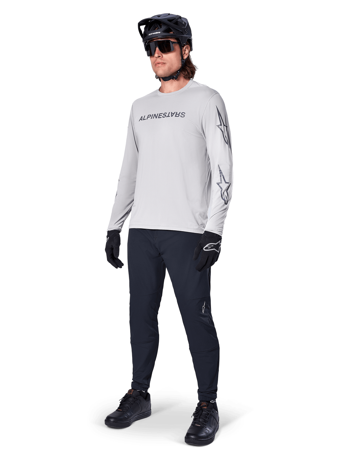 Alpinestars A-Dura Switch Long Sleeve Jersey, jasnoszara, noszona przez rowerzystę z czarnymi spodniami MTB i kaskiem, czarny logotyp na piersi i grafiki gwiazd na rękawach, profesjonalna odzież do jazdy terenowej