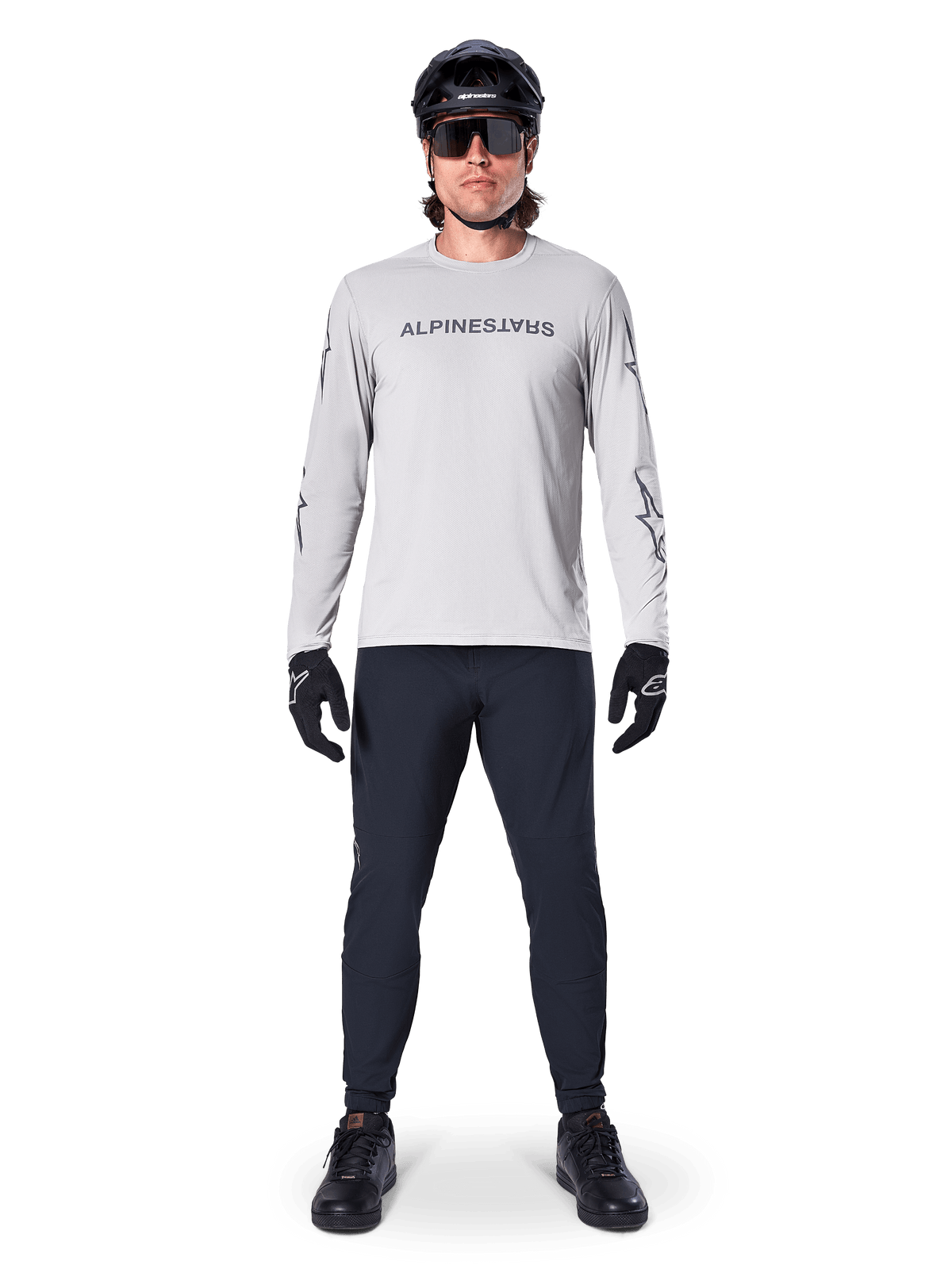 Koszulka Alpinestars A-Dura Switch z długim rękawem w kolorze jasnoszarym, ze stylizowanym logotypem na piersi, noszona z czarnymi spodniami, kaskiem i rękawiczkami, profesjonalna odzież do kolarstwa górskiego