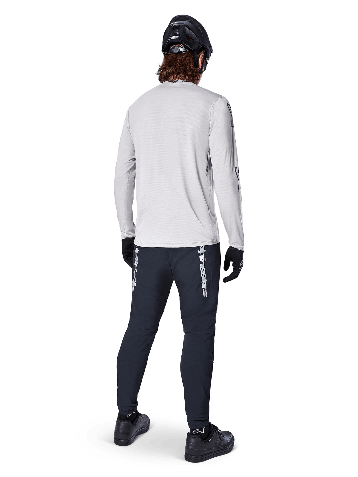 Alpinestars A-Dura Switch Long Sleeve Jersey, jasnoszara koszulka rowerowa, widok z tyłu na rowerzystę w czarnych spodniach, kasku i rękawicach, odzież MTB o wysokiej wydajności z wydłużonym tyłem