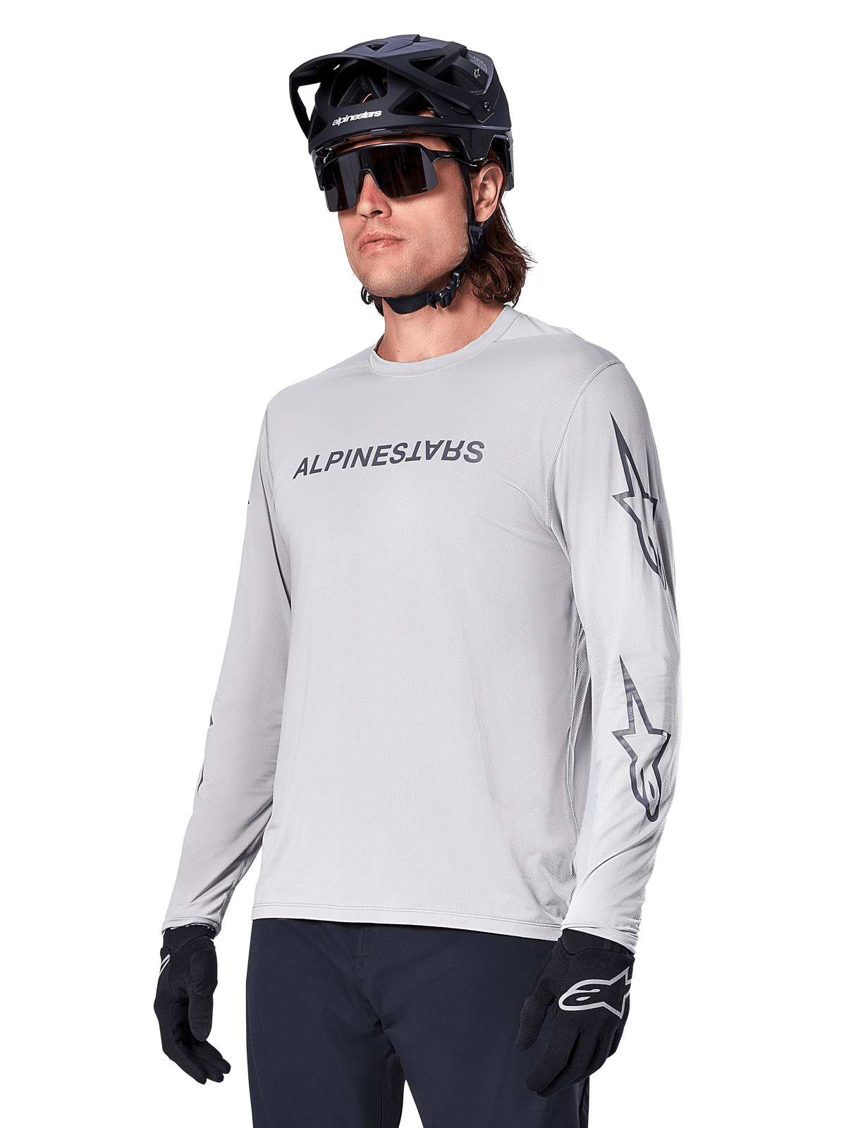 Alpinestars A-Dura Switch Long Sleeve Jersey, koszulka na rower górski, jasnoszara, czarny nadruk z nazwą marki na piersi i logo z gwiazdą na rękawach, noszona z czarnym kaskiem rowerowym MTB, ciemnymi okularami i czarnymi rękawiczkami, sportowa odzież do kolarstwa terenowego