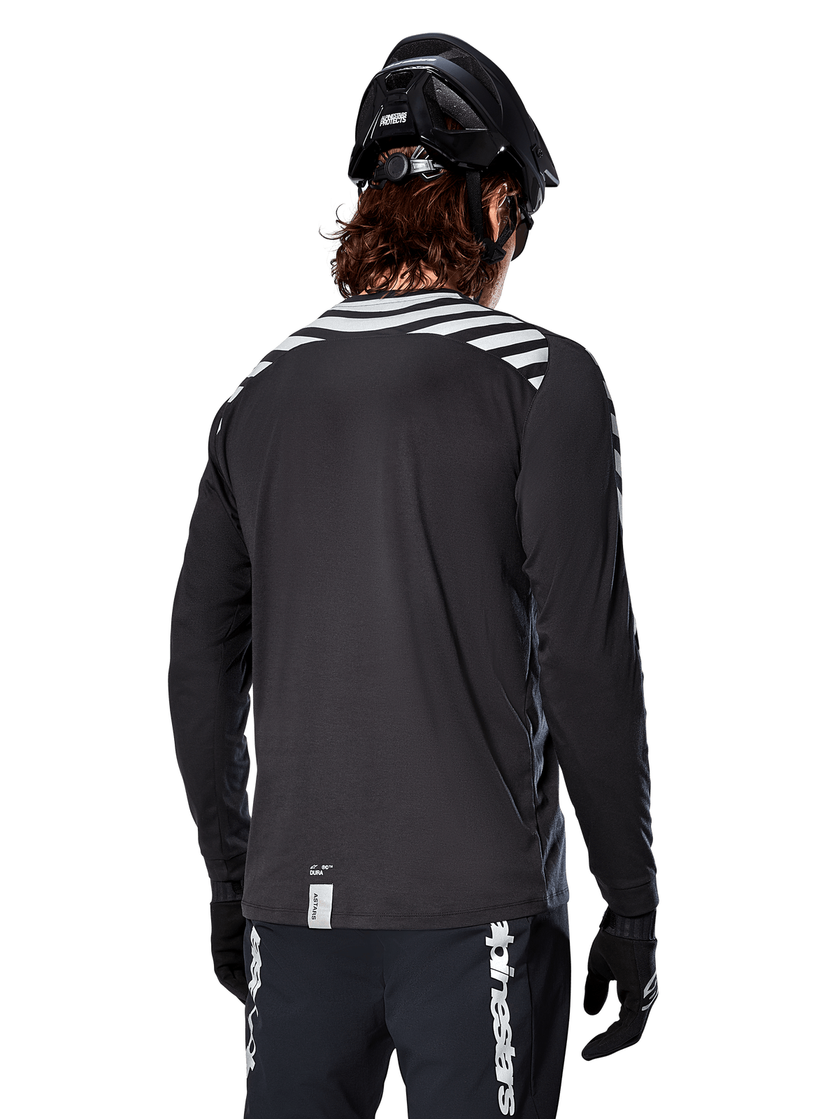 Alpinestars A-Dura Dri Oscar Long Sleeve Jersey, Black Reflective, rear view showing reflective diagonal stripes on shoulders and a longer drop tail, worn with black MTB helmet and gloves, technical gear for mountain biking