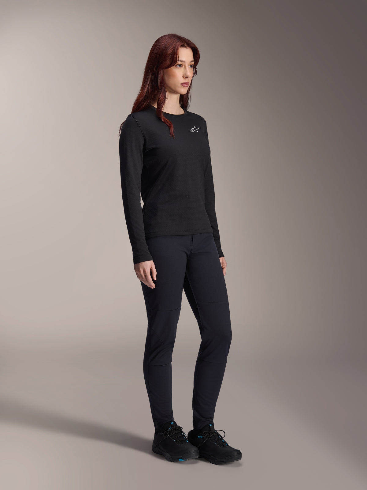 Kobieta ubrana w koszulkę Alpinestars Stella A-Dura Thermal, czarna koszulka MTB z długim rękawem i białym logo na piersi, zestawiona z dopasowanymi czarnymi spodniami i butami sportowymi, portret studyjny profesjonalnej odzieży rowerowej
