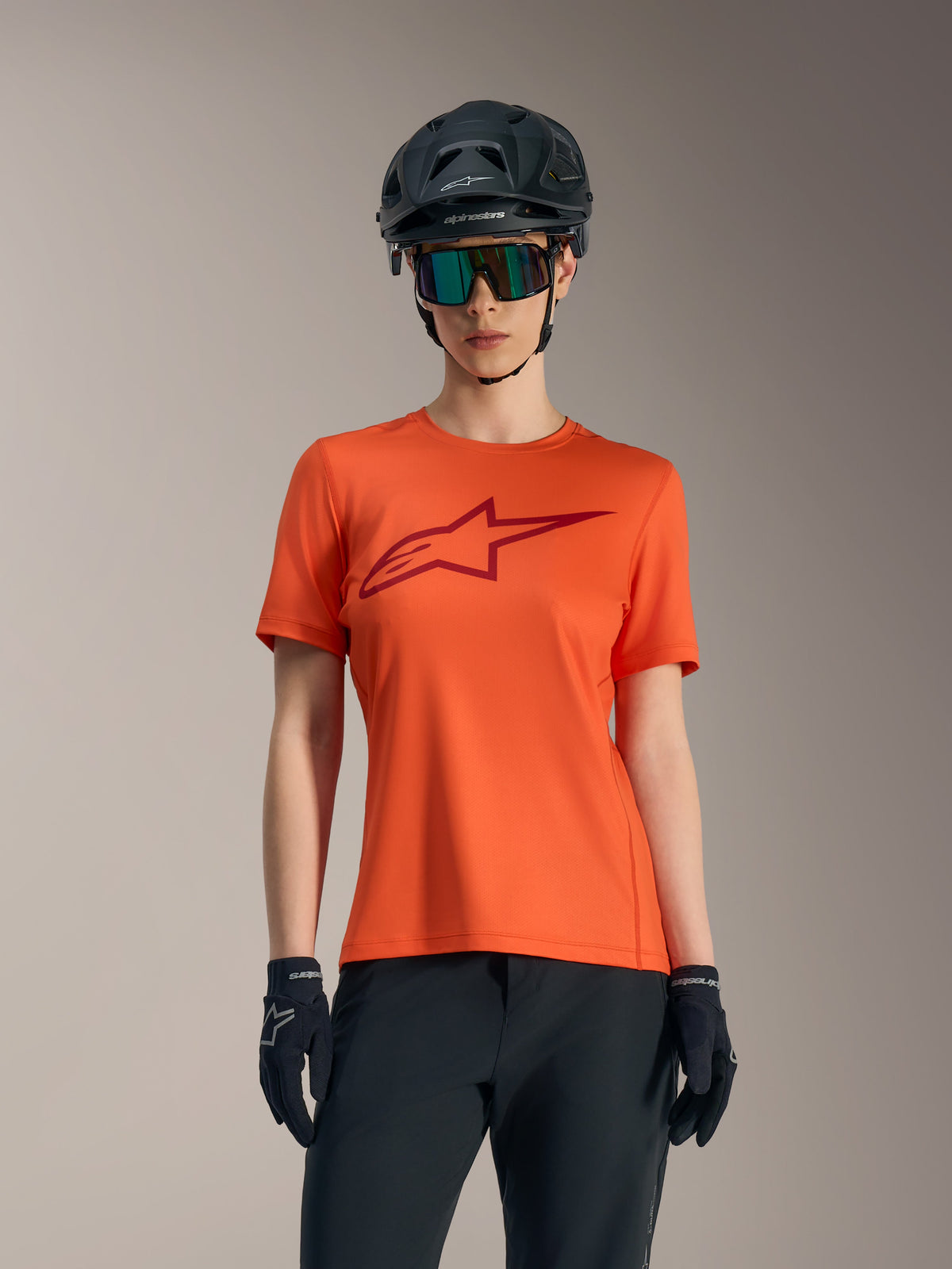 Koszulka Alpinestars Stella A-Dura Ride, pomarańczowa z czerwonym logo, czarny kask MTB i okulary przeciwsłoneczne.