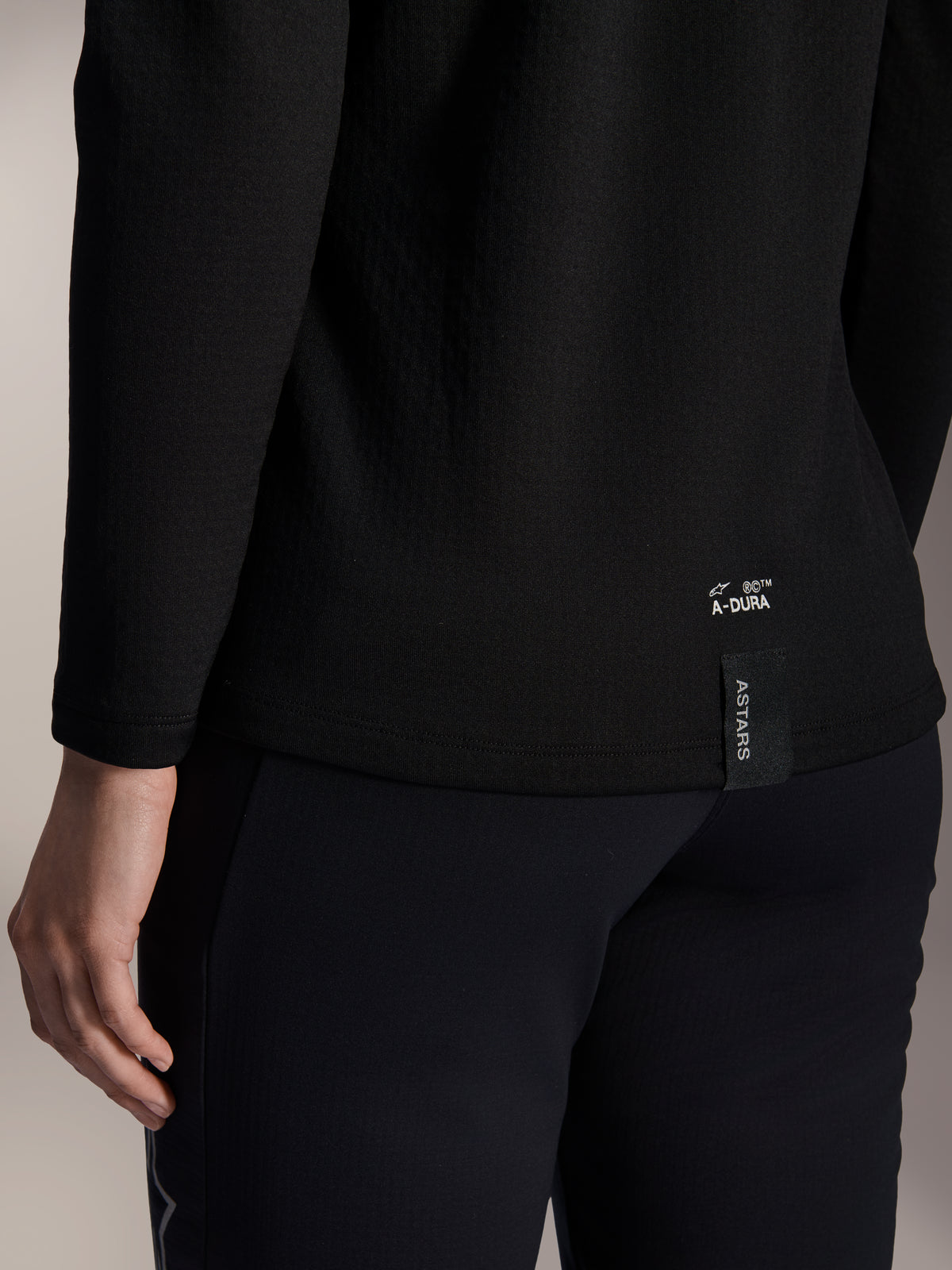 Alpinestars Stella A-Dura Thermal Jersey, czarna damska koszulka termiczna MTB, zbliżenie na dolne wykończenie tyłu z białym brandingiem A-DURA i pionową metką z logo ASTARS, zaprojektowana do jazdy na rowerze górskim w chłodne dni