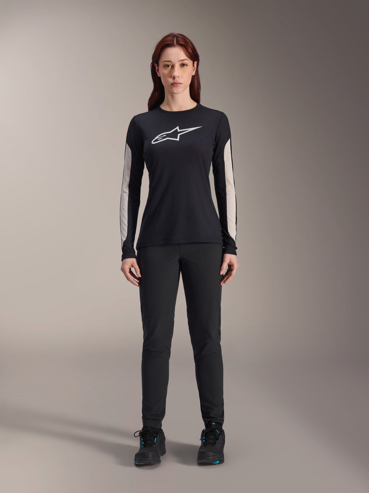Kobieta w koszulce Alpinestars Stella A-Dura Dri Astar Long Sleeve Jersey, czarna z białym logo na piersi i jasnymi panelami z siatki na rękawach, w zestawieniu z czarnymi spodniami i butami, techniczna odzież rowerowa MTB