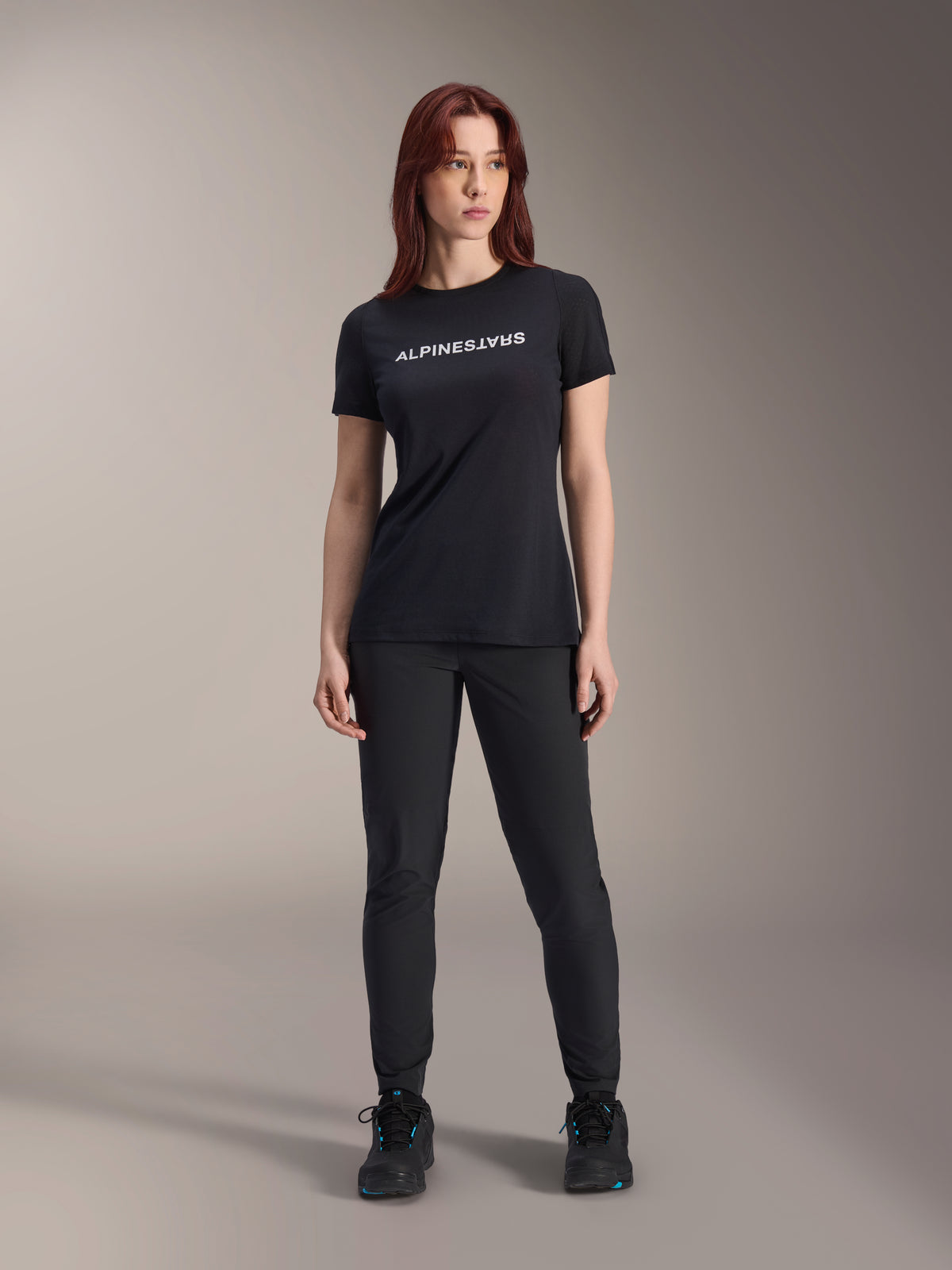 Modelka w damskiej koszulce Alpinestars Stella A-Dura Dri Switch z krótkim rękawem w kolorze czarnym, z białym logo na piersi, czarne spodnie techniczne i czarne buty sportowe, ujęcie studyjne całej sylwetki