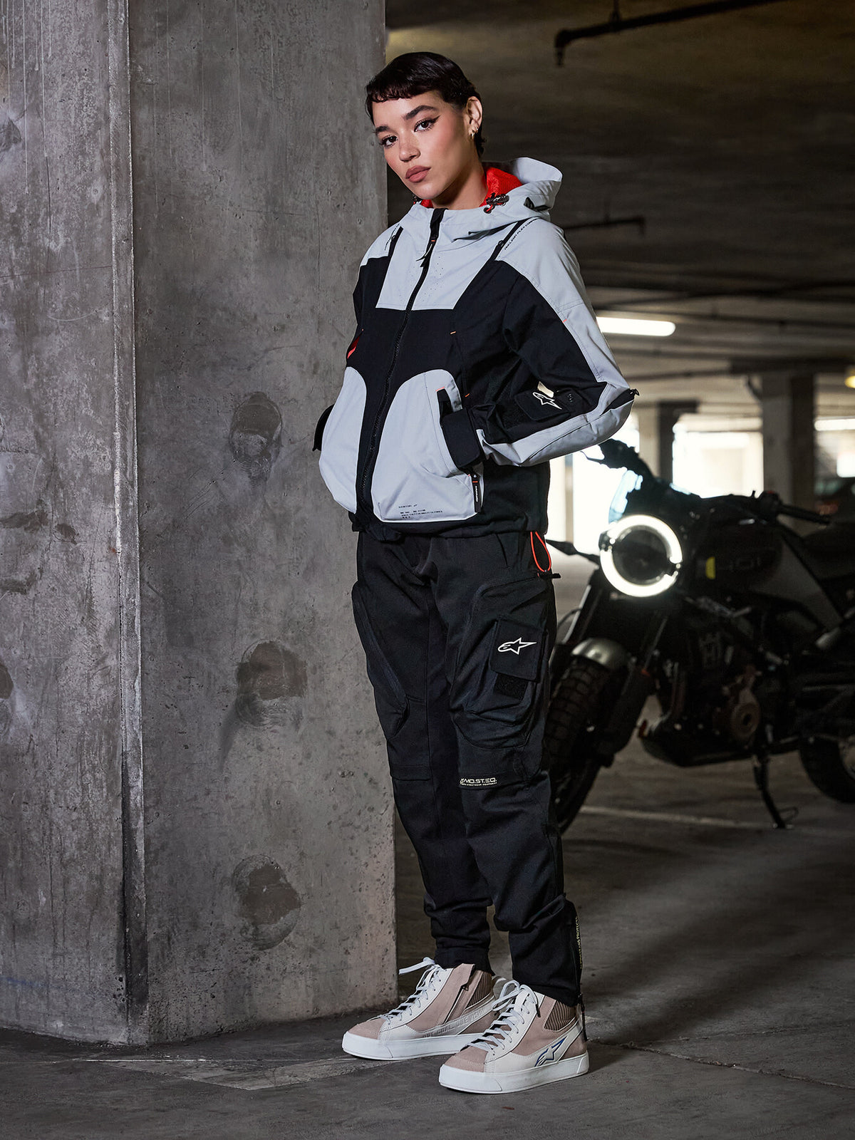 Spodnie damskie Alpinestars Actea, czarne, motocyklistka w motocyklowych spodniach typu cargo, szaro-czarnej kurtce technicznej i beżowych sneakersach, w otoczeniu miejskiego garażu parkingowego z motocyklem.