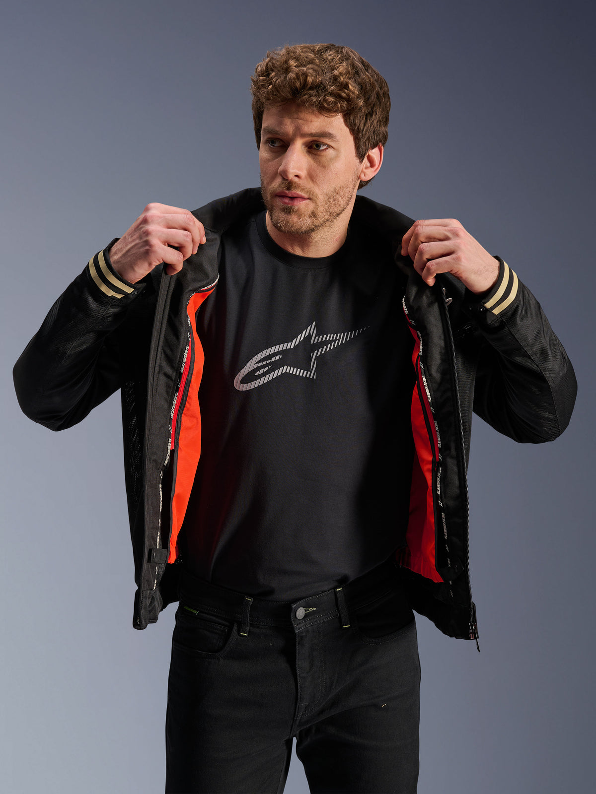 Mężczyzna w czarnej kurtce Alpinestars Ignite Air z siatkowym korpusem i jaskrawopomarańczową odpinaną wiatroodporną podszewką, czarny t-shirt z logo Astars, czarne spodnie, współczesny miejski styl motocyklowy