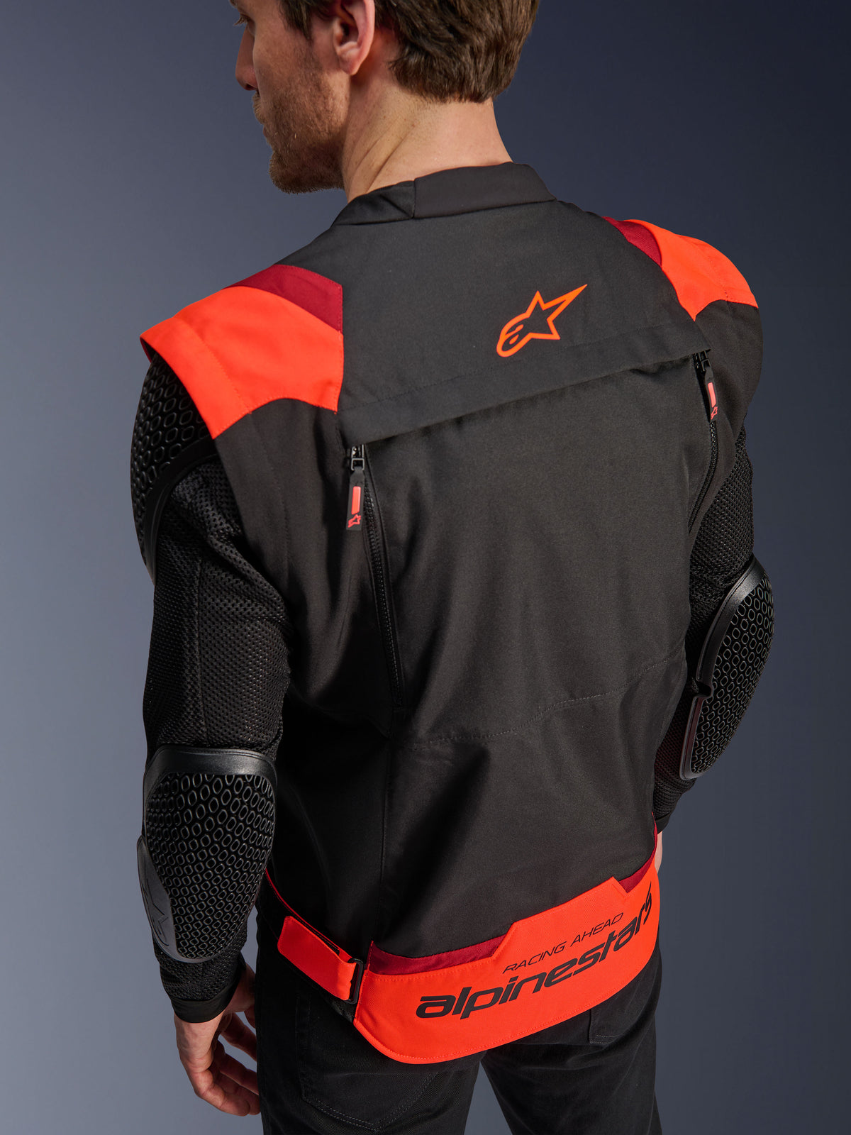 Kurtka Alpinestars T-Stunt Air, czarna z fluorescencyjną i ciemną czerwienią, widok z tyłu na modelu, siatkowe rękawy z ochraniaczami łokci o strukturze plastra miodu, logo gwiazdy Alpinestars na górze pleców, branding „RACING AHEAD” w dolnej części, modułowy sportowy krój.
