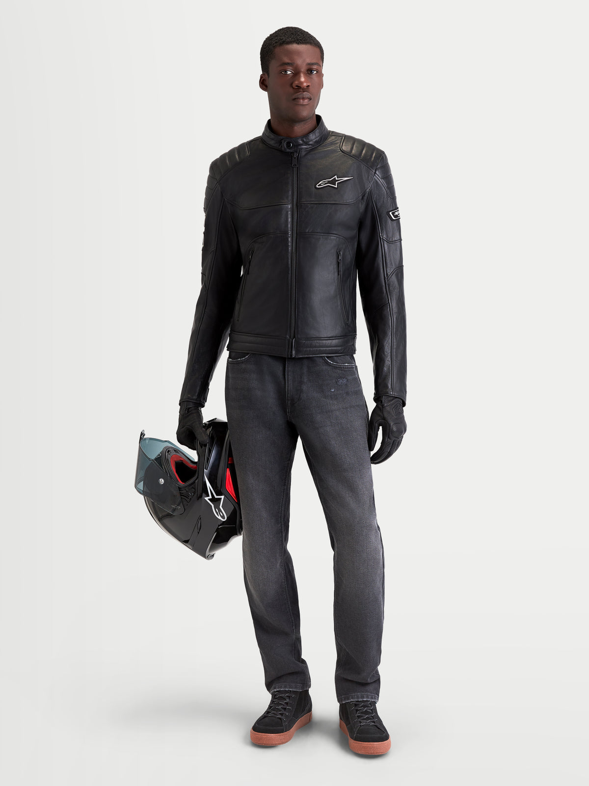 Alpinestars Revern Kurtka skórzana, męska urbanistyczna motocyklowa kurtka skórzana w kolorze czarnym, widok z przodu na modela w ciemnych jeansach trzymającego kask, z konstrukcją z postarzanej skóry bydlęcej i klasycznym motocyklowym stylem