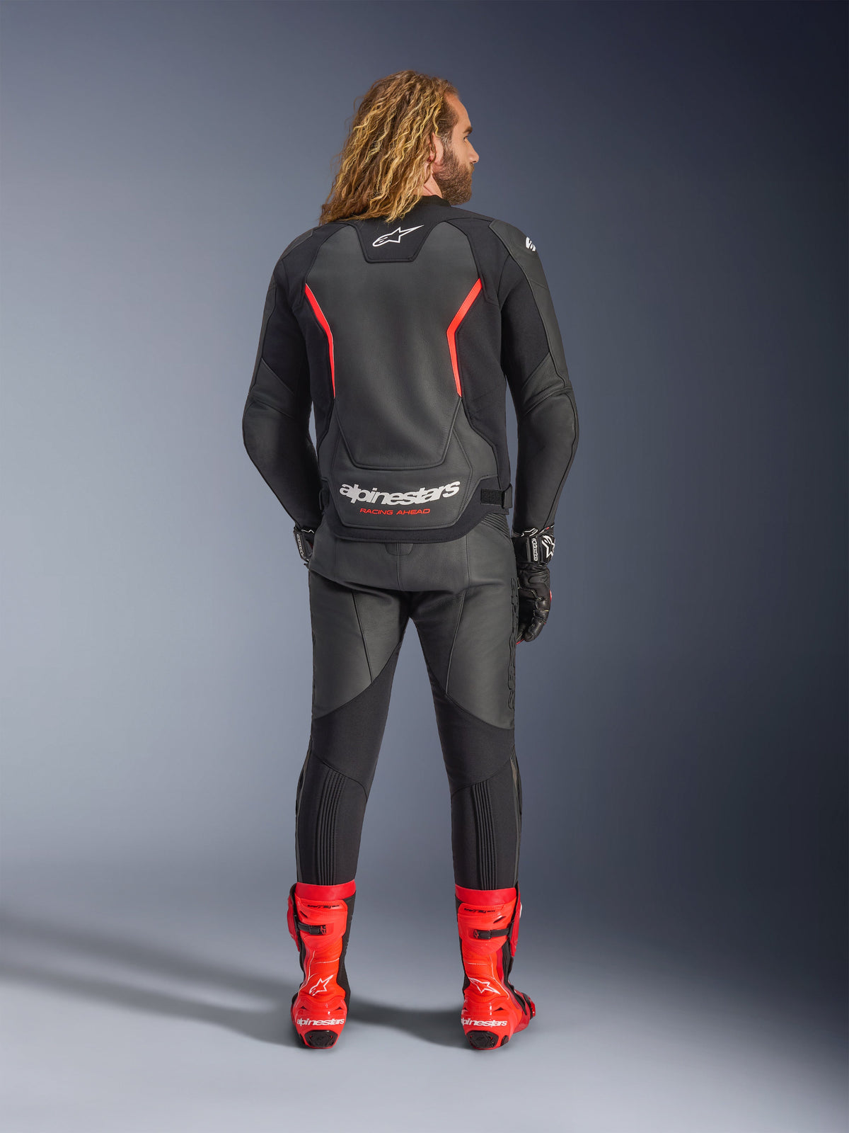 Skórzana kurtka Alpinestars GP Force V2, czarno-czerwona Fluo, widziana z tyłu, noszona z dopasowanymi czarnymi spodniami skórzanymi i jaskrawoczerwonymi butami wyścigowymi, z czerwonymi liniami akcentowymi i wyraźnym białym logo Alpinestars na plecach, profesjonalny sportowy strój motocyklowy