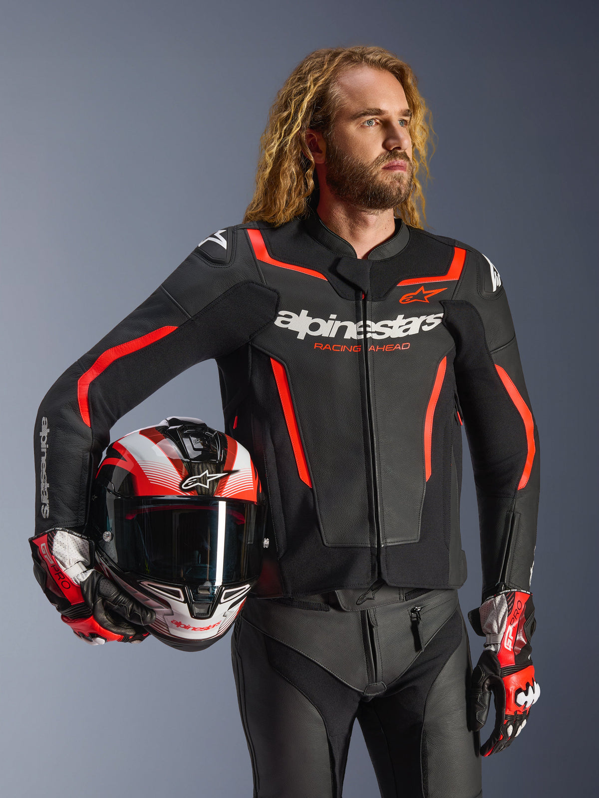 Skórzana kurtka Alpinestars GP Force V2, czarno-czerwona fluo, noszona przez modela trzymającego pasujący kask, białe logo Alpinestars i czerwone akcenty, w zestawieniu ze skórzanymi spodniami i rękawicami wyścigowymi, sportowa odzież motocyklowa na drogę i tor