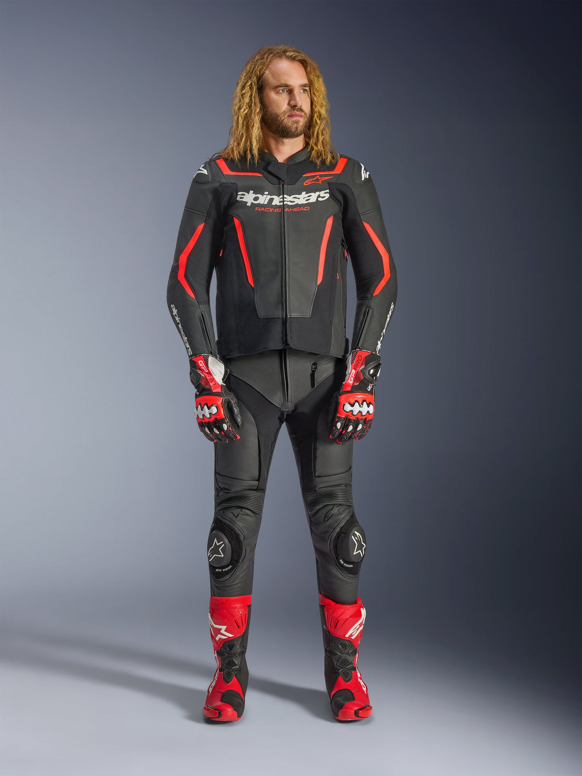 Męski model w skórzanej kurtce Alpinestars GP Force V2 w kolorze czarno-czerwonym Fluo, zestawionej z dopasowanymi skórzanymi spodniami, czerwonymi rękawicami wyścigowymi i czerwonymi butami sportowymi, profesjonalna odzież motocyklowa