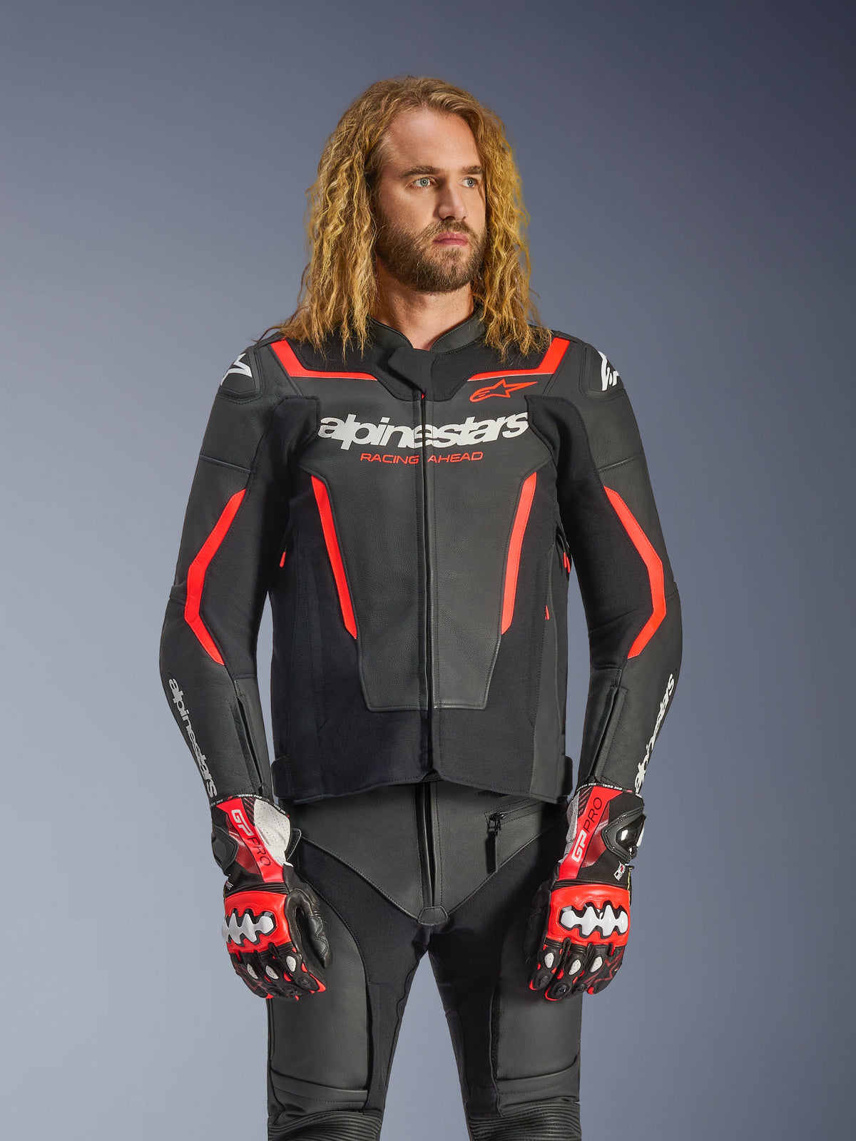 Alpinestars GP Force V2 Leather Jacket, sportowa kurtka motocyklowa, czarno-czerwona Fluo, prezentowana przez modela z długimi falowanymi włosami, białe logo Alpinestars i napis „RACING AHEAD”, w zestawie z dopasowanymi skórzanymi spodniami i rękawicami, profesjonalna odzież motocyklowa