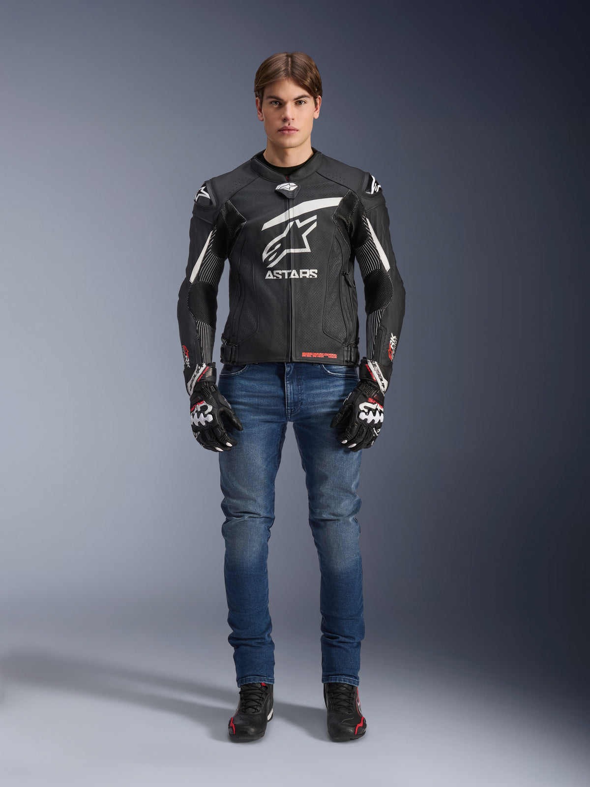Męski model w skórzanej kurtce Alpinestars GP Plus R V4 Rideknit w kolorze czarno-białym, stylizowany z niebieskimi dżinsami motocyklowymi, czarnymi rękawicami i butami do jazdy, z dużym białym logo Astars na piersi, perforowanymi panelami skórzanymi i białymi pasami wyścigowymi na rękawach, widok całej sylwetki stojącej