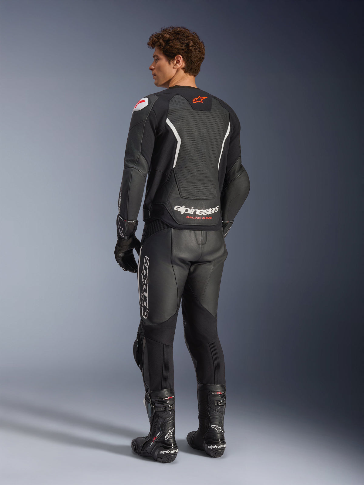 Alpinestars GP Force V2 Air Leather Jacket, czarno-biała, widok z tyłu na modelu z perforowanym panelem tylnym dla wentylacji, białe logo i czerwona gwiazda, noszona z pasującymi skórzanymi spodniami wyścigowymi, rękawicami i butami