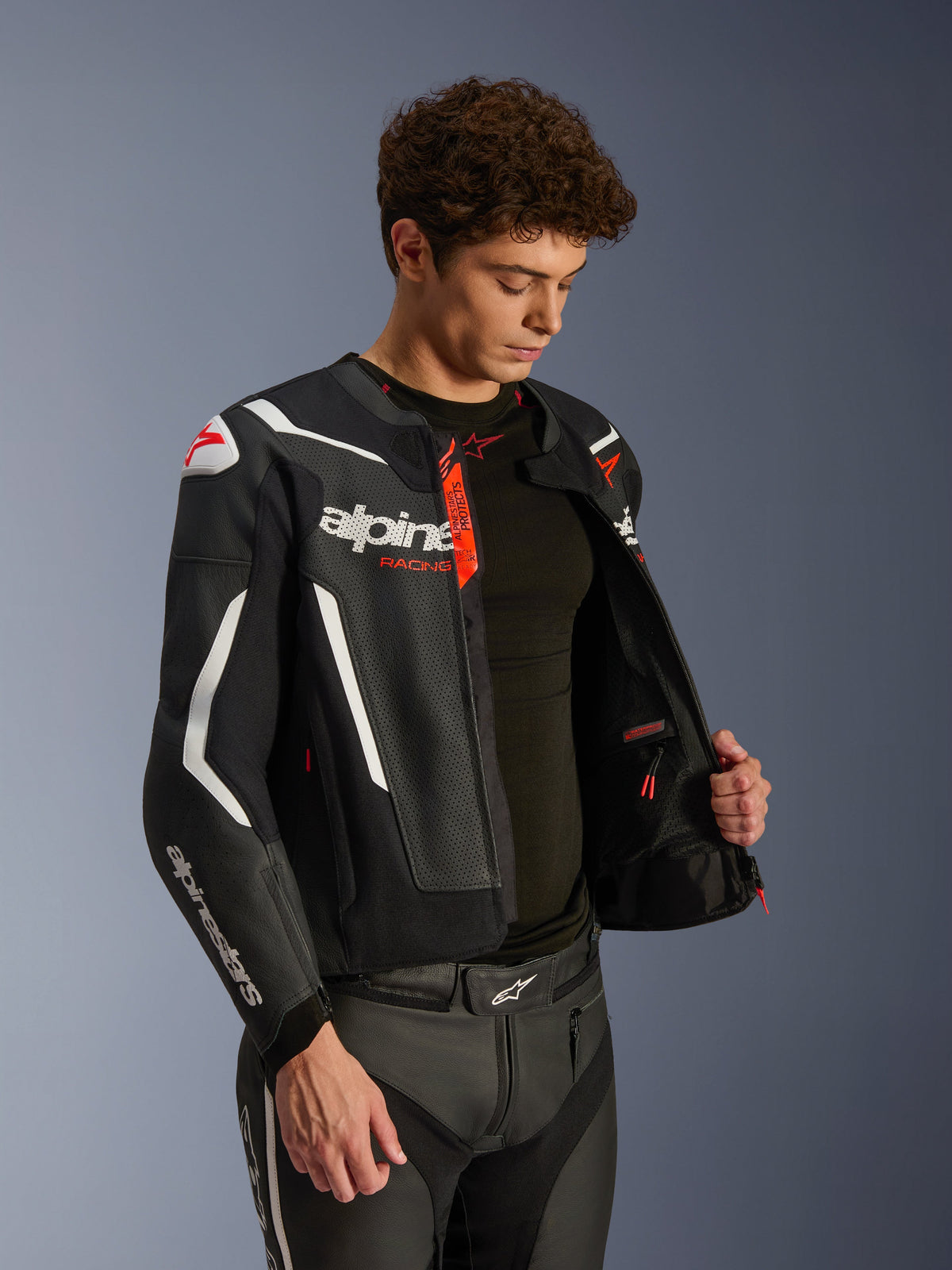 Kurtka skórzana Alpinestars GP Force V2 Air, czarno-biała z czerwonymi akcentami, konstrukcja z perforowanej skóry, rozpięta, pokazująca wnętrze i czarną warstwę bazową, zestawiona z pasującymi skórzanymi spodniami, profesjonalne zdjęcie studyjne odzieży motocyklowej