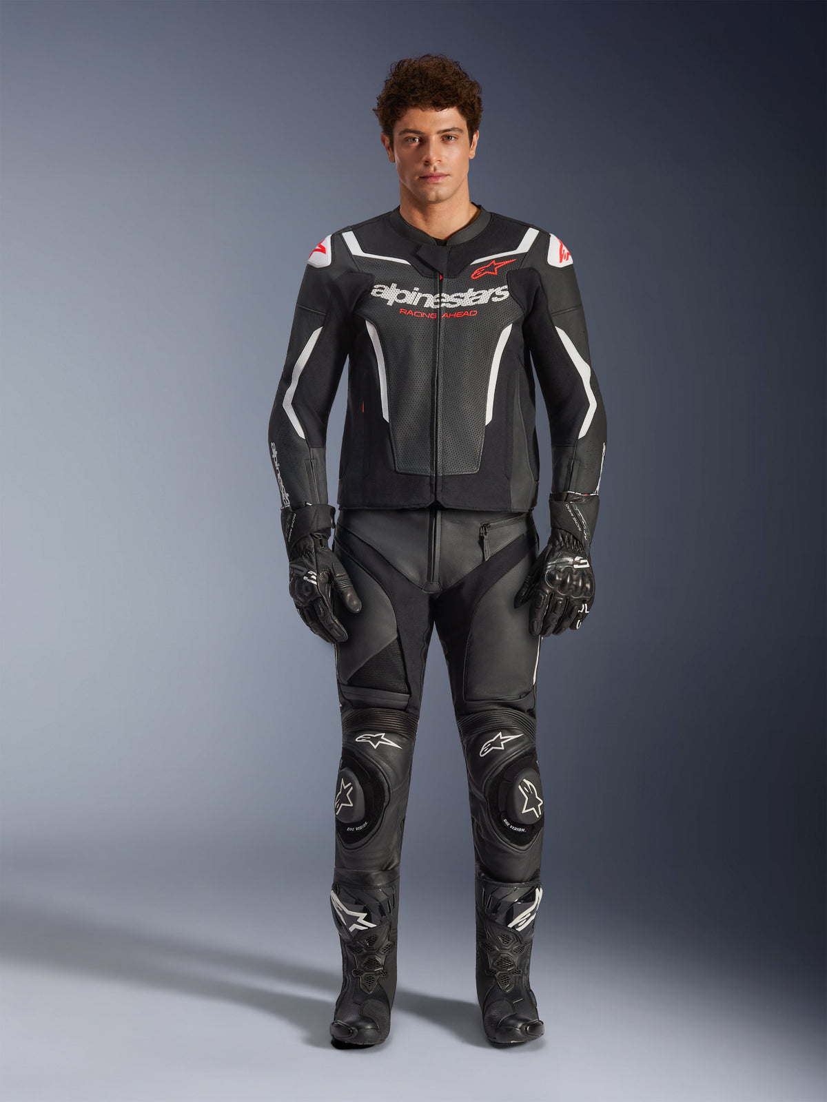 Męski model w kurtce skórzanej Alpinestars GP Force V2 Air, czarno-biała, w zestawie z pasującymi skórzanymi spodniami, rękawicami i butami, sportowy widok z przodu, perforowana skórzana konstrukcja z białymi akcentami i logo Alpinestars