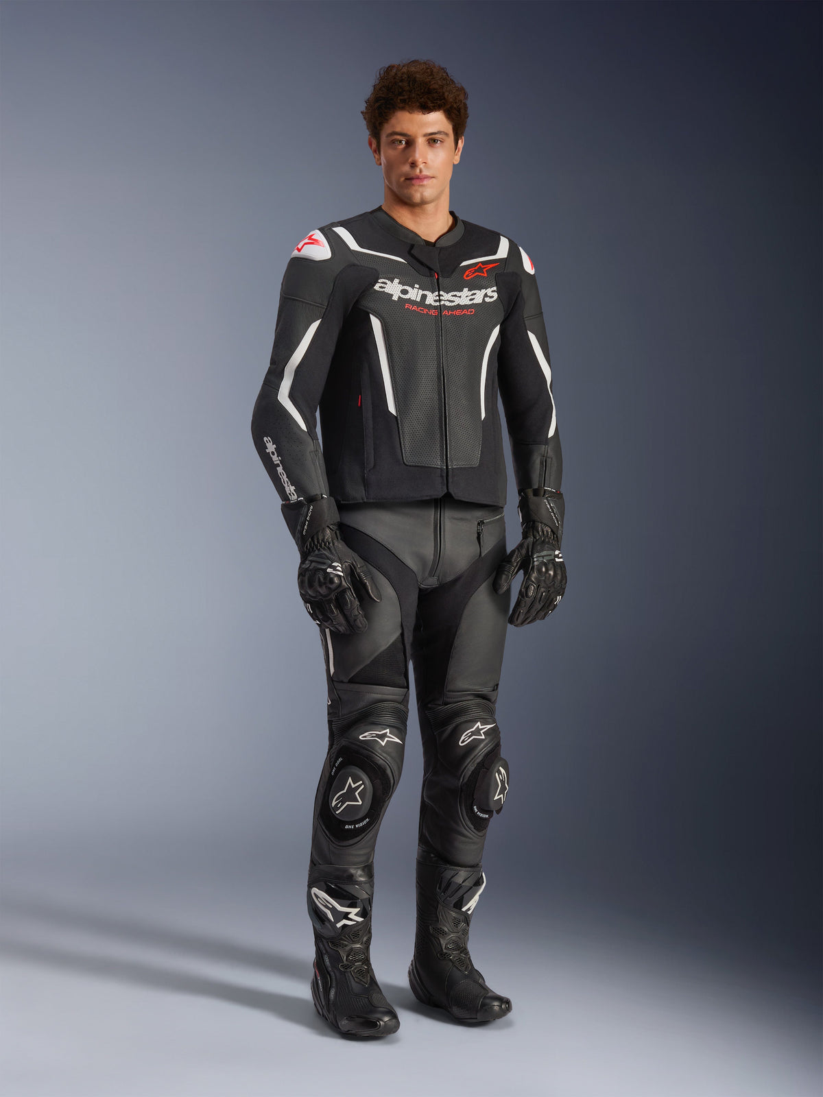Kurtka skórzana Alpinestars GP Force V2 Air w kolorze czarno-białym, prezentowana przez motocyklistę z dopasowanymi czarnymi spodniami wyścigowymi, rękawicami i butami, wyposażona w perforowane panele skórzane, białe akcenty i czerwone logo Alpinestars na klatce piersiowej, sportowa odzież motocyklowa