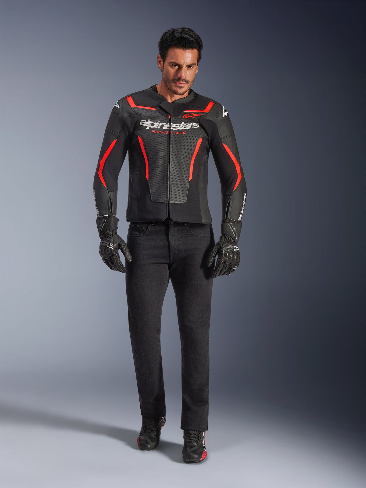 Motocyklista w skórzanej kurtce Alpinestars GP Force V2 Air, czarno-czerwona Fluo, zestawiona z czarnymi jeansami, rękawicami i butami, perforowana skórzana konstrukcja, białe logo i sportowe czerwone akcenty