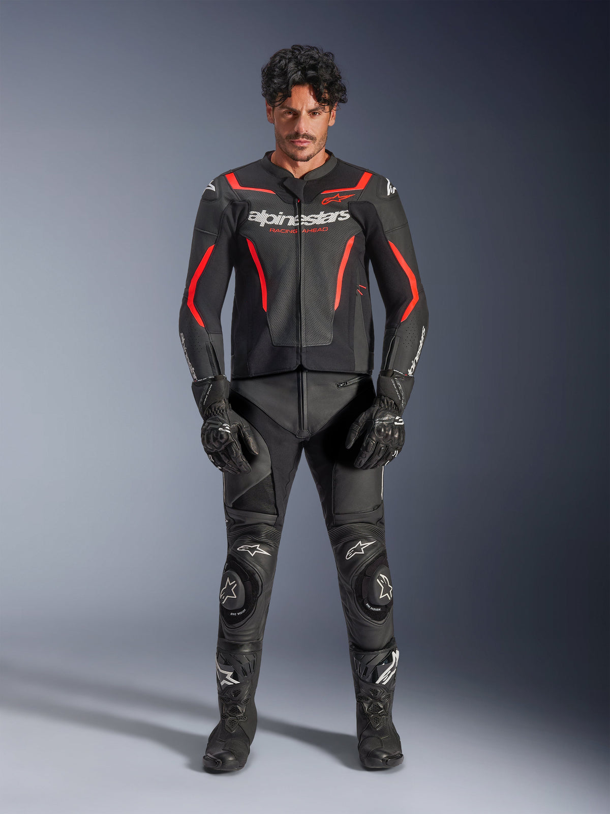 Męski model w skórzanej kurtce Alpinestars GP Force V2 Air w kolorze czarno-czerwonym (Black Red Fluo), z perforowanymi panelami na klatce piersiowej i białym logo, w zestawie z czarnymi skórzanymi spodniami motocyklowymi, czarnymi rękawicami i czarnymi butami, tworzący kompletny wyścigowy look.
