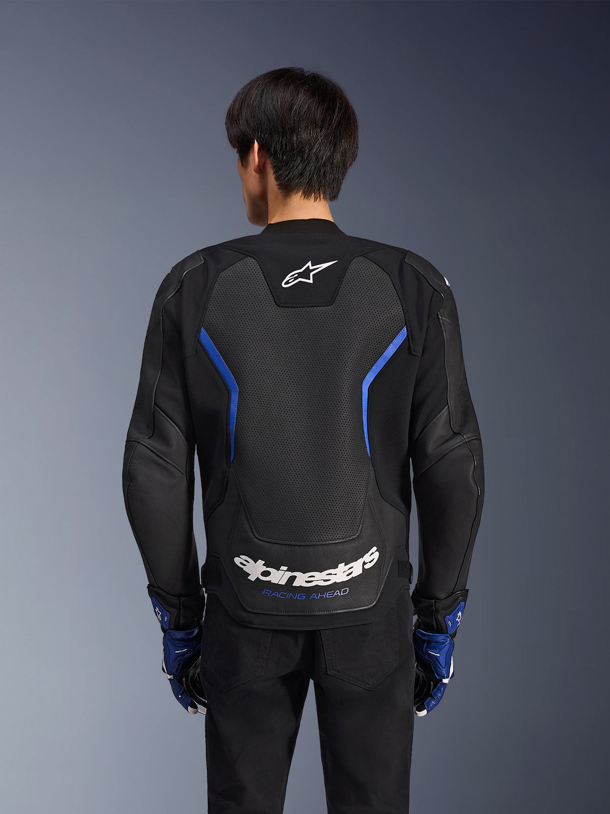 Alpinestars GP Force V2 Airflow Leather Jacket, skórzana kurtka motocyklowa, czarna z metalicznymi niebieskimi akcentami, widok z tyłu, duży perforowany panel na plecach dla wentylacji, białe logo Alpinestars, noszona z niebieskimi rękawicami i czarnymi spodniami do jazdy sportowej