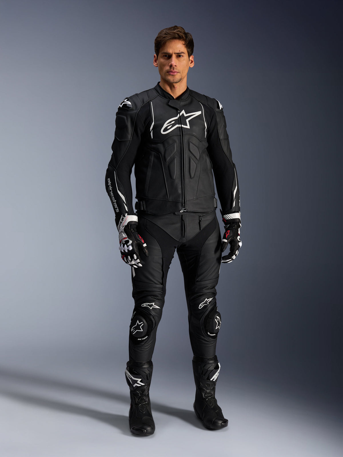 Alpinestars Missile V3 Leather Pants, czarne spodnie skórzane, noszone przez motocyklistę jako element kompletnego stroju wyścigowego, białe logo Alpinestars, ochronne ślizgacze kolan, buty wyścigowe i rękawice