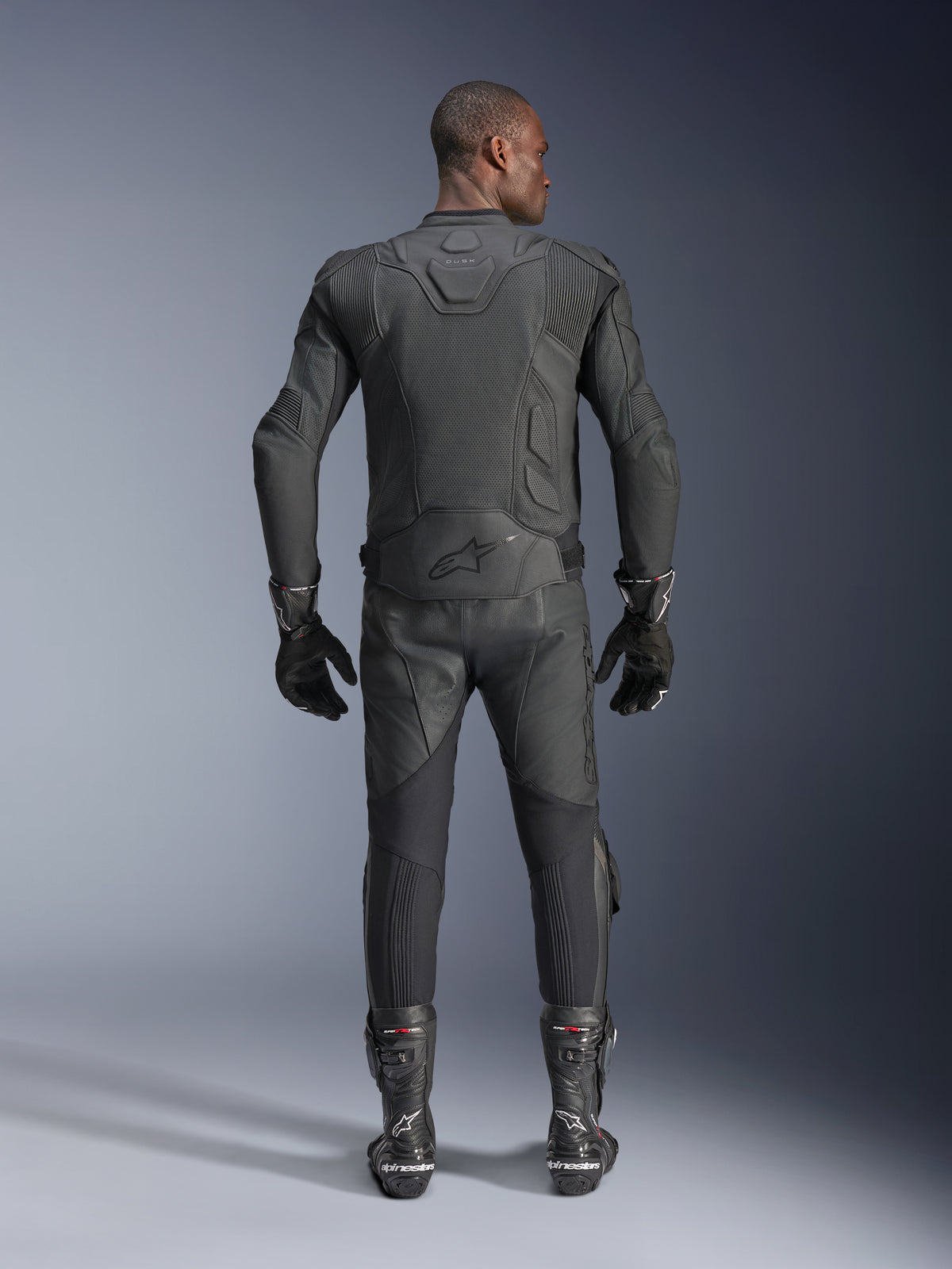 Male model shown from the back wearing the Alpinestars Dusk Airflow Leather Jacket in Black, paired with matching black leather racing pants, gloves, and boots, featuring extensive perforations for ventilation and the Alpinestars logo on the lower back.