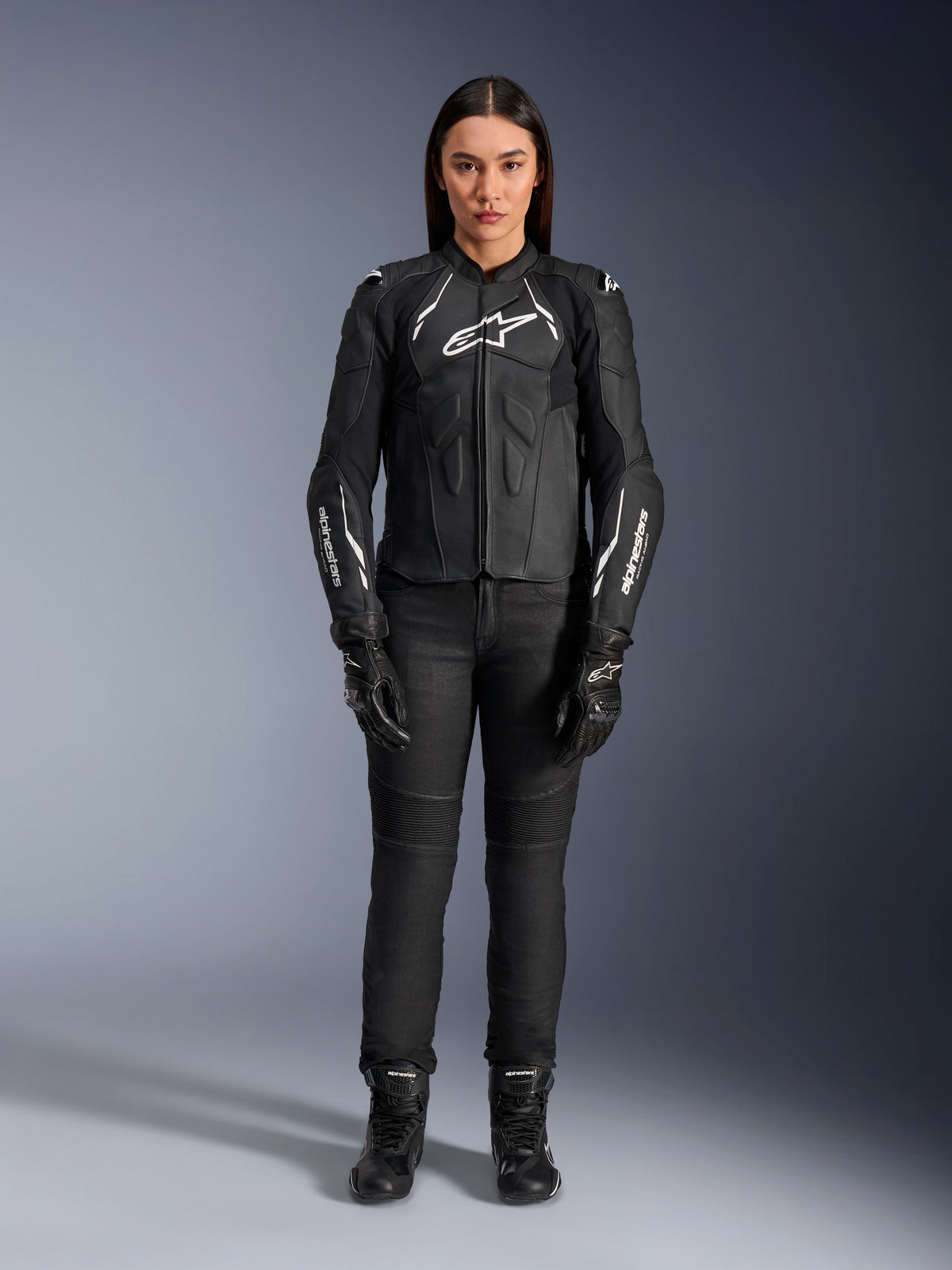 Damska skórzana kurtka Alpinestars Stella Dusk, czarno-biała, prezentowana przez modelkę z dopasowanymi czarnymi spodniami i butami, widok z przodu, z białym logo Alpinestars na klatce piersiowej i rękawach, najwyższej jakości odzież ochronna z bydlęcej skóry dla kobiet