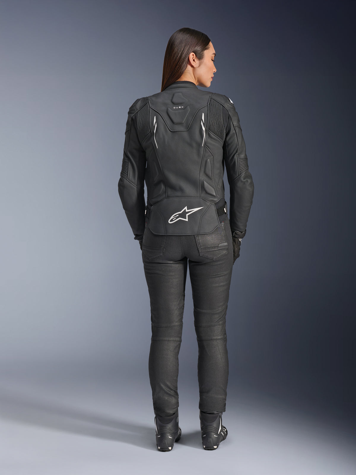 Damska skórzana kurtka Alpinestars Stella Dusk, czarna, widok z tyłu z białym logo Alpinestars na dole pleców i brandingiem „DUSK” w górnej części pleców, noszona z czarnymi spodniami motocyklowymi i butami, prezentująca dopasowany damski krój oraz ochronną skórzaną konstrukcję.