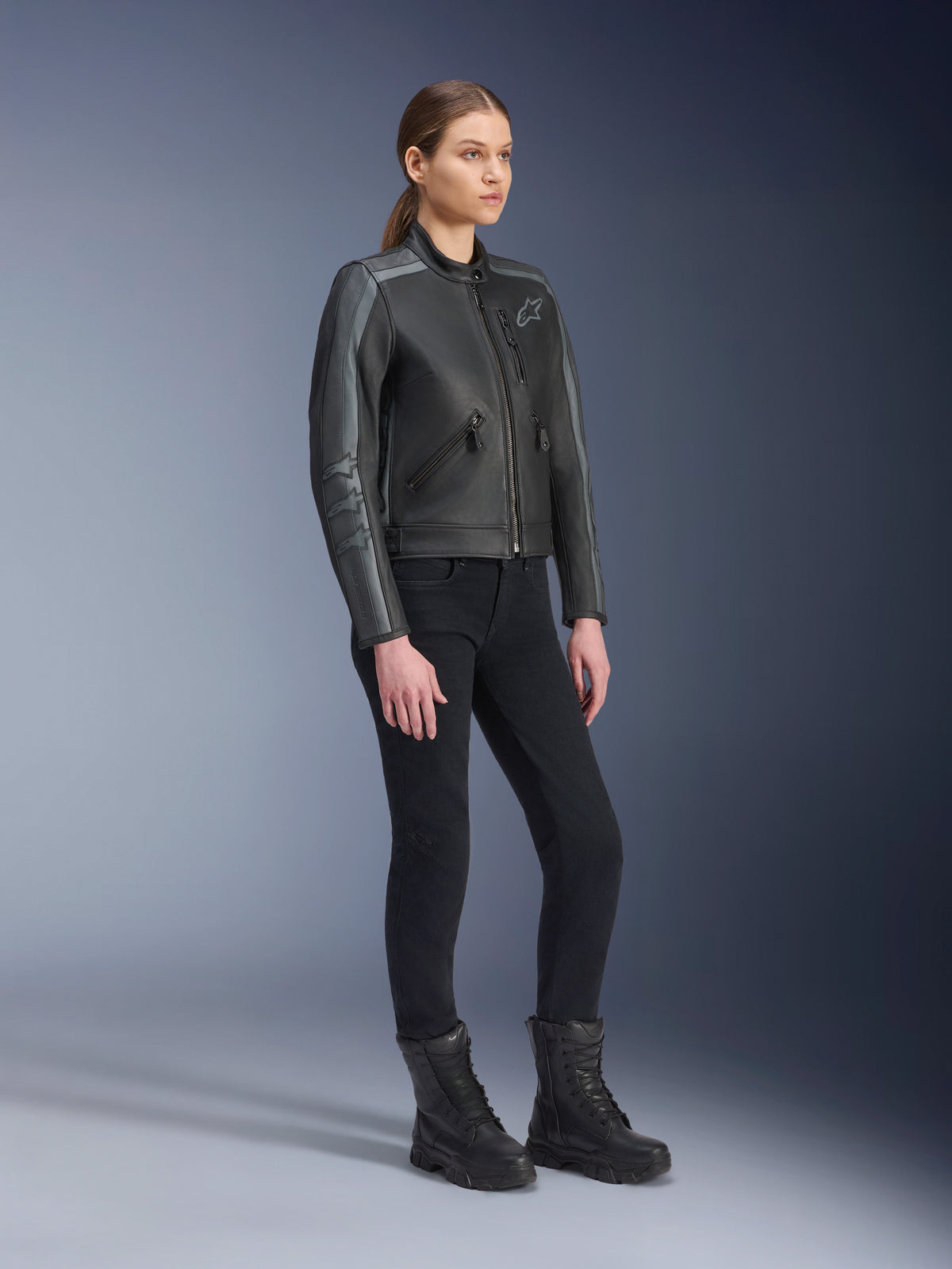 Alpinestars Stella Dyno Leather Jacket, damska kurtka motocyklowa, czarna i ciemnoszara, noszona z czarnymi spodniami i butami motocyklowymi, z szarymi wyścigowymi pasami na rękawach i skórzanym wzornictwem typu heritage