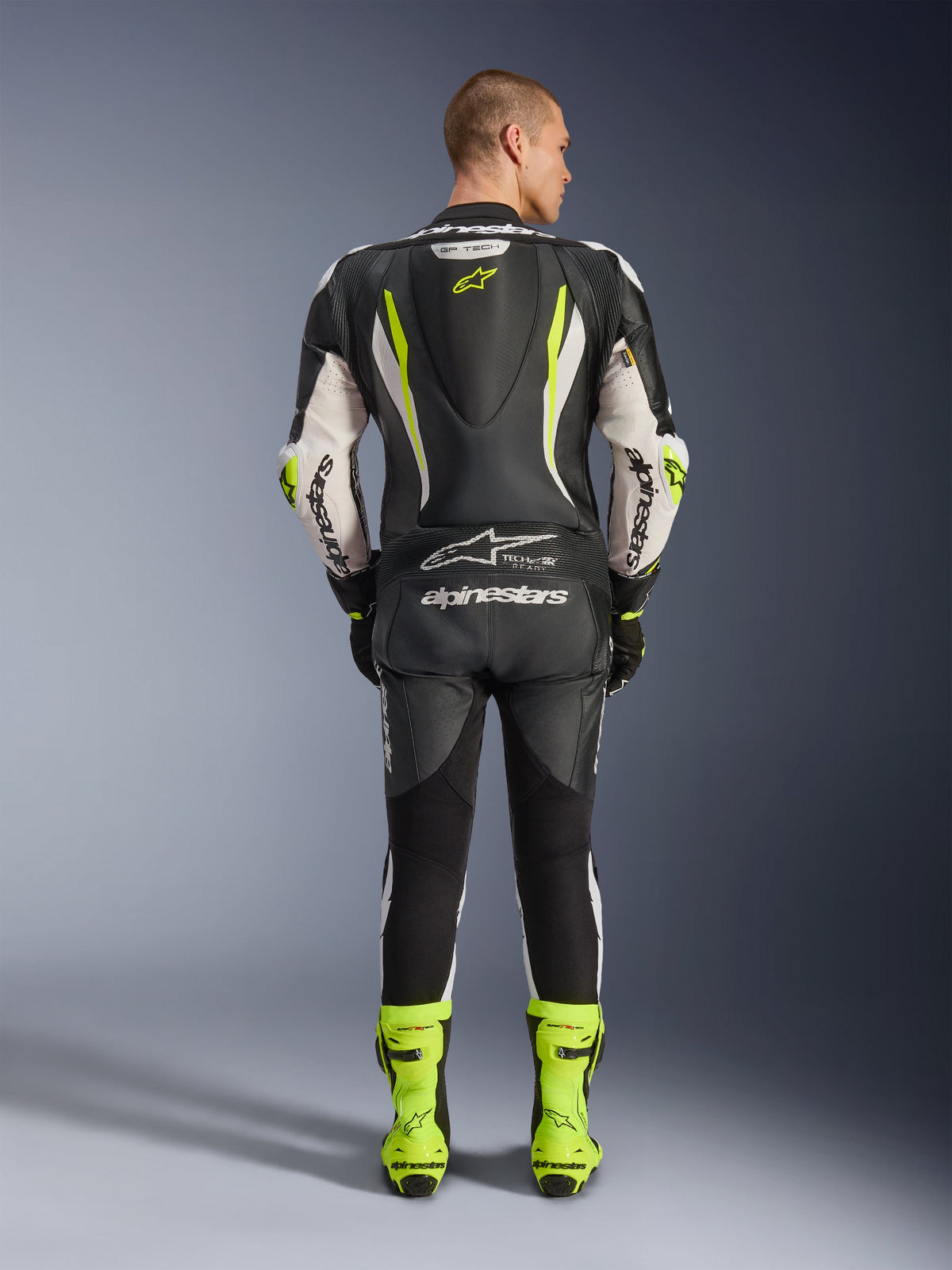 Jednoczęściowy kombinezon skórzany Alpinestars GP Tech V5, widok z tyłu, czarno-biało-żółty fluo, aerodynamiczny garb z żółtym logo gwiazdy, białe logo Alpinestars na rękawach i plecach, pasujące neonowe żółte buty wyścigowe, profesjonalny sprzęt motocyklowy na tor