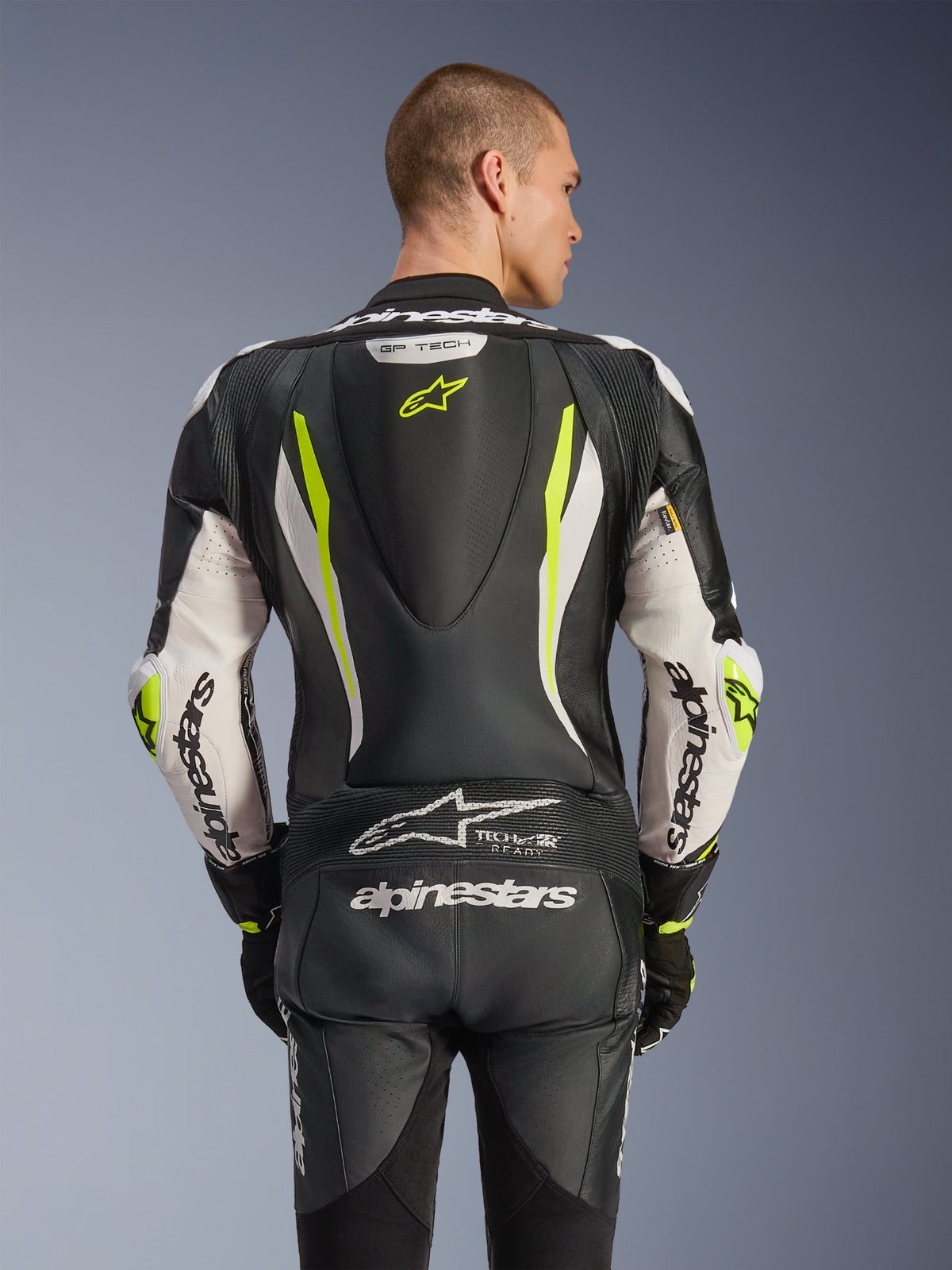 Alpinestars GP Tech V5 One Piece Leather Suit, widok z tyłu na modelu, kolor czarno-biały i żółty fluo, z aerodynamicznym garbem, dużym logo Alpinestars na rękawach i siedzeniu, elastycznymi panelami akordeonowymi oraz logo Tech-Air Ready