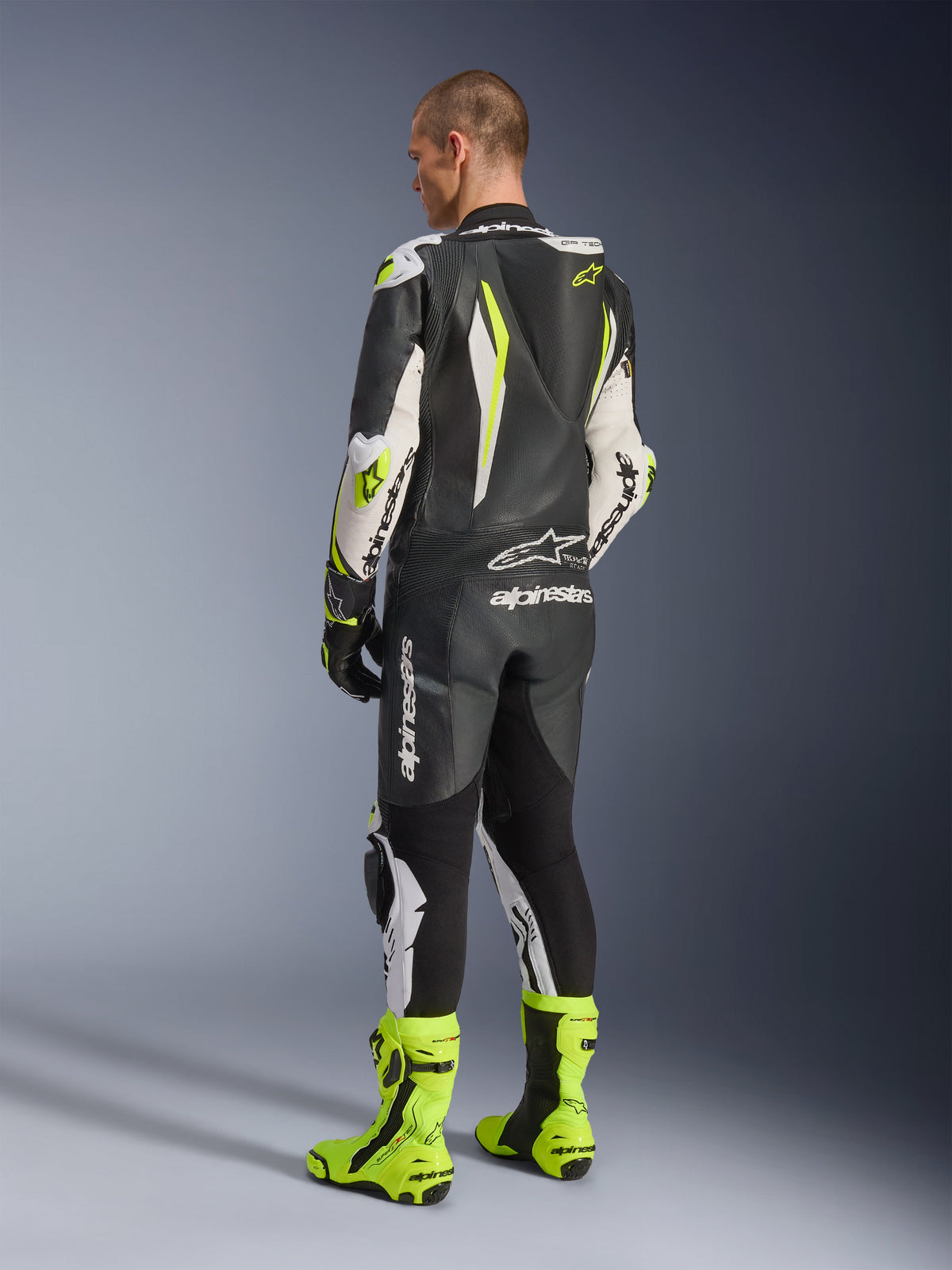 Jednoczęściowy kombinezon skórzany GP Tech V5, czarno-biało-żółty fluo, widok od tyłu motocyklisty w pełnym kombinezonie wyścigowym z pasującymi żółtymi butami fluo, posiada aerodynamiczny garb i wyraźne logo Alpinestars, profesjonalna odzież motocyklowa