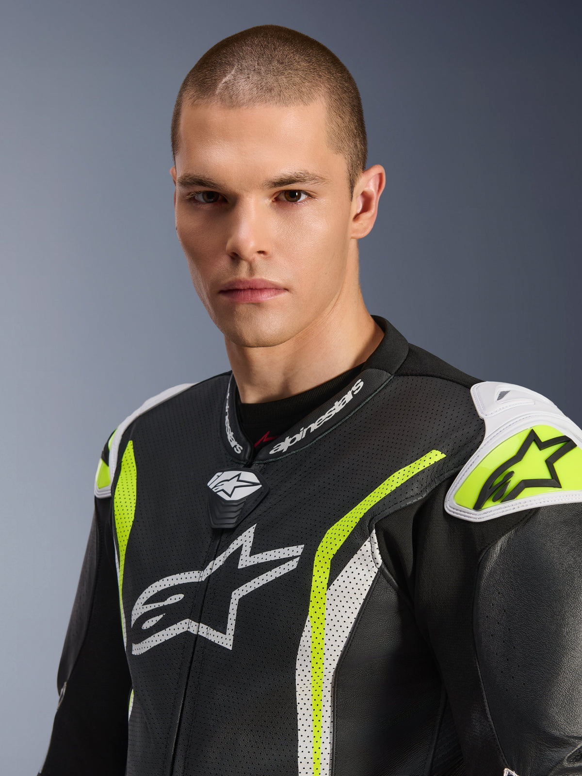Alpinestars GP Tech V5 One Piece Leather Suit, zbliżenie na modela w czarno-biało-żółtym kombinezonie fluo, perforowana skóra, białe logo Alpinestars na klatce piersiowej i ramionach, jaskrawe żółte akcenty, profesjonalna odzież wyścigowa