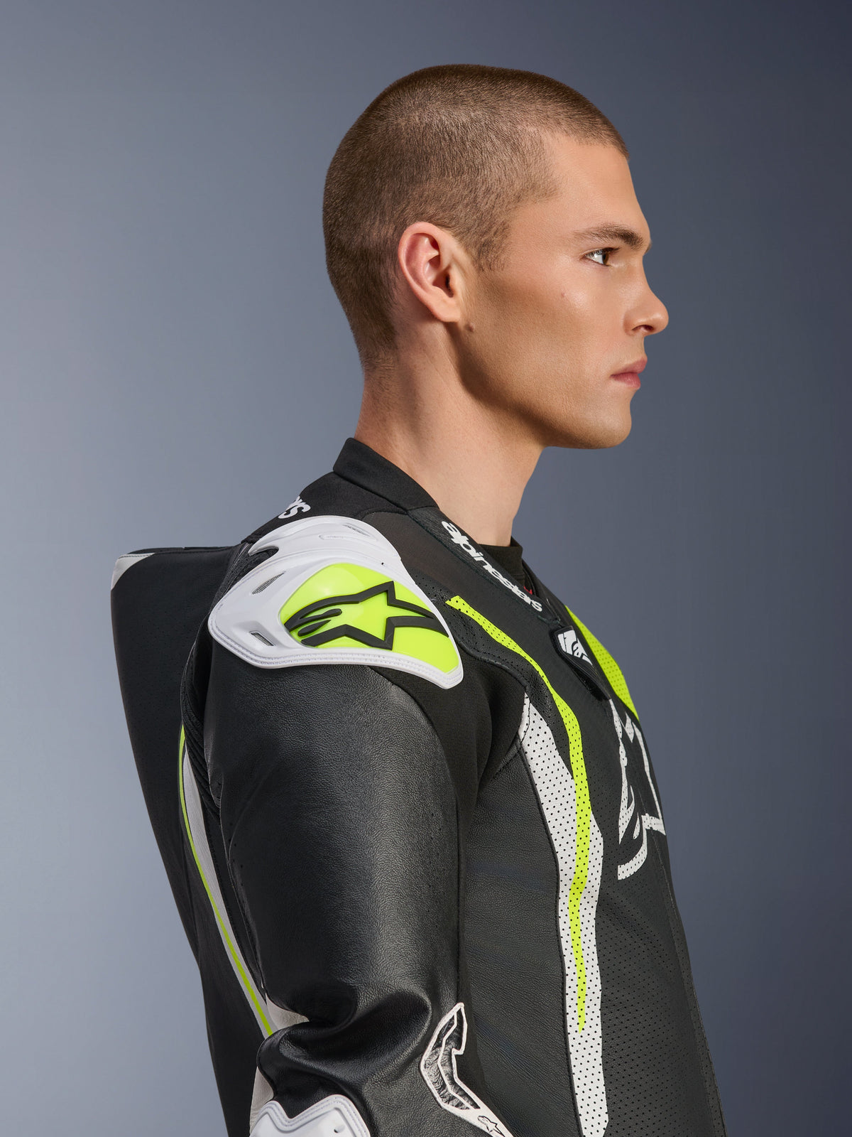 Alpinestars GP Tech V5 One Piece Leather Suit, męski model w jednoczęściowym kombinezonie skórzanym w kolorze Black White Yellow Fluo, widok z profilu podkreślający aerodynamiczny garb i białe slidery barkowe, wyścigowa odzież motocyklowa z detalami z perforowanej skóry