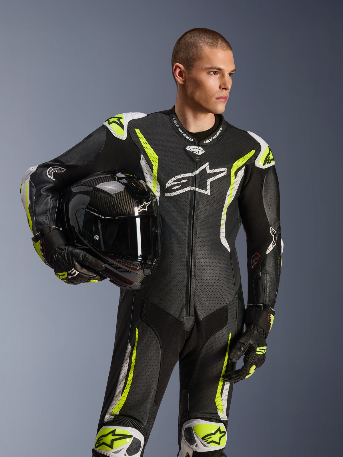 Alpinestars GP Tech V5 One Piece Leather Suit, czarny, biały i żółty fluo, męski model trzymający czarny kask motocyklowy, perforowane panele, białe i fluorescencyjne żółte akcenty, wyraźne logo Alpinestars, profesjonalna odzież wyścigowa