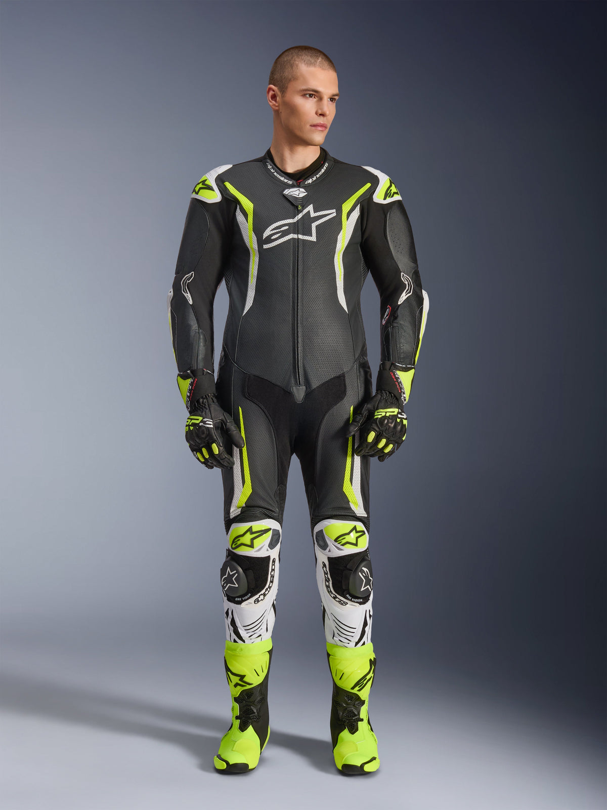 Alpinestars GP Tech V5 One Piece Leather Suit, profesjonalny motocyklowy kombinezon wyścigowy w kolorze czarnym i neonowo-żółtym fluo z białymi akcentami, noszony przez modela z dopasowanymi rękawicami i butami wyścigowymi, o aerodynamicznym designie i z ochronnymi sliderami.
